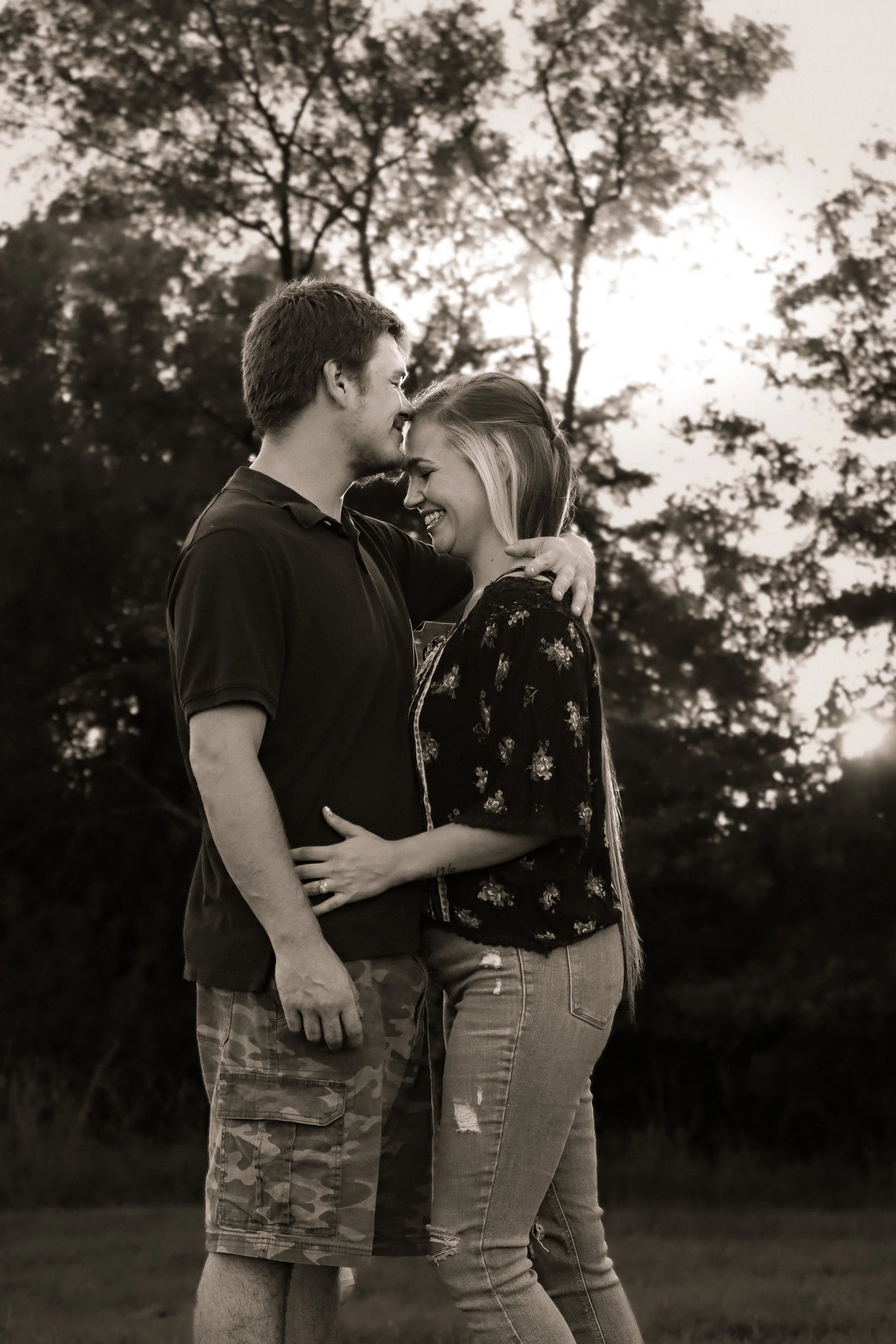 A smiling couple embraces outdoors during sunset, with trees and the sky in the background.