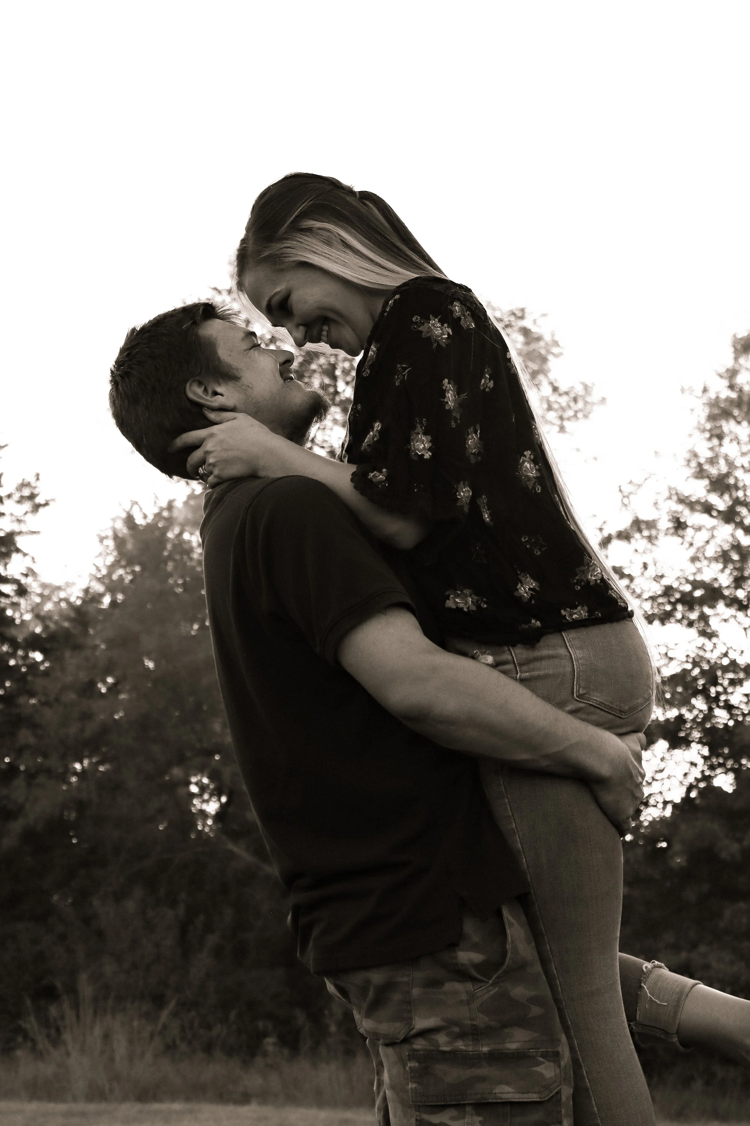 A couple outdoors, with the man lifting the woman in his arms as they smile and look at each other, background of trees, black and white.