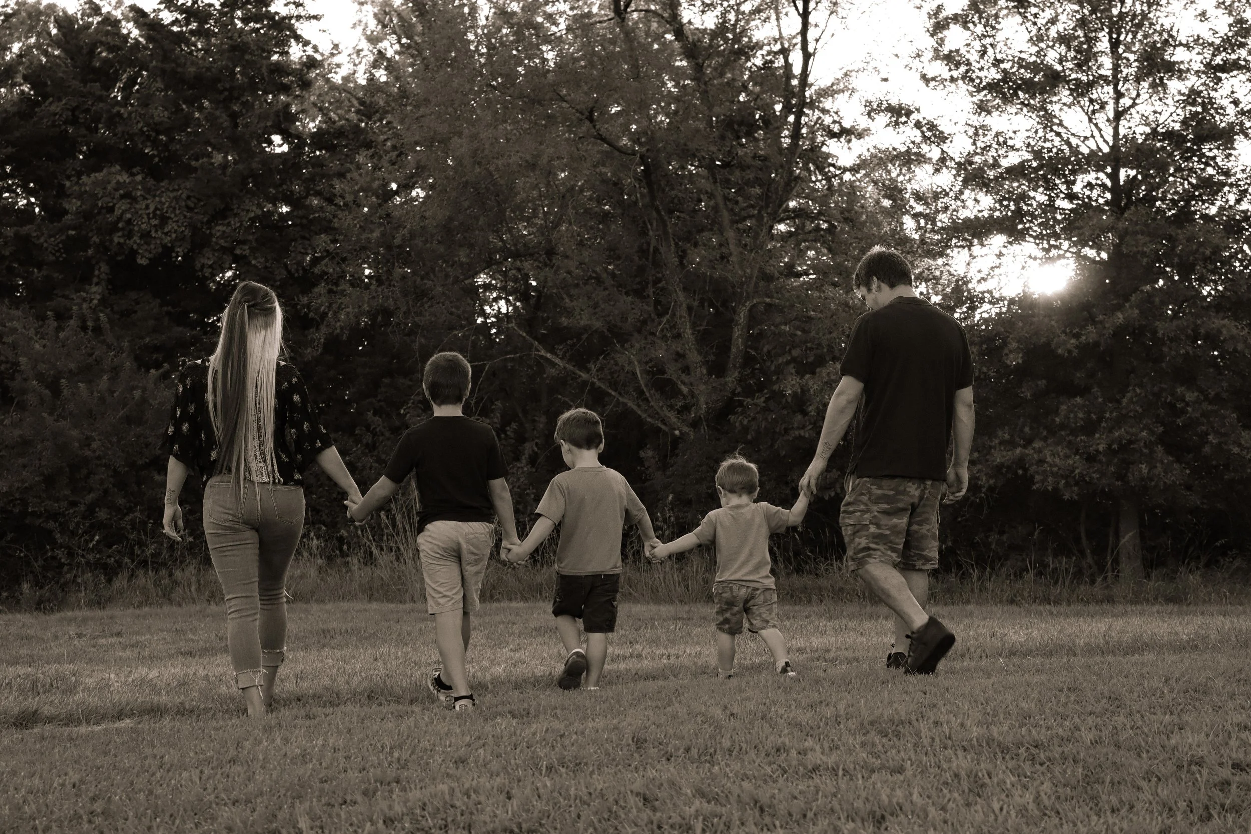 A family of six holding hands and walking in a grassy field during sunset, with trees in the background, in black and white.