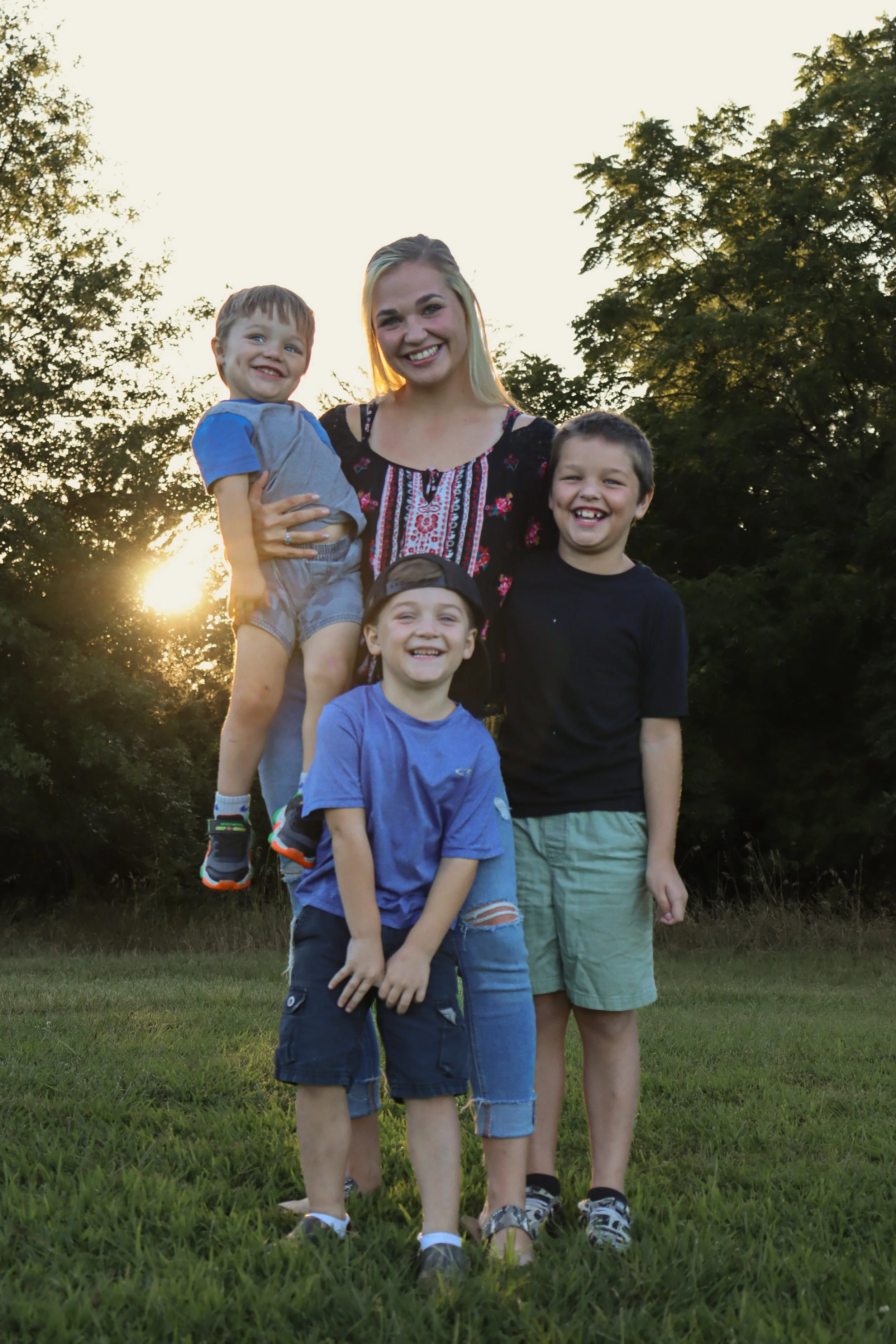 A woman with four young boys outdoors at sunset, standing on grassy field with trees in background.