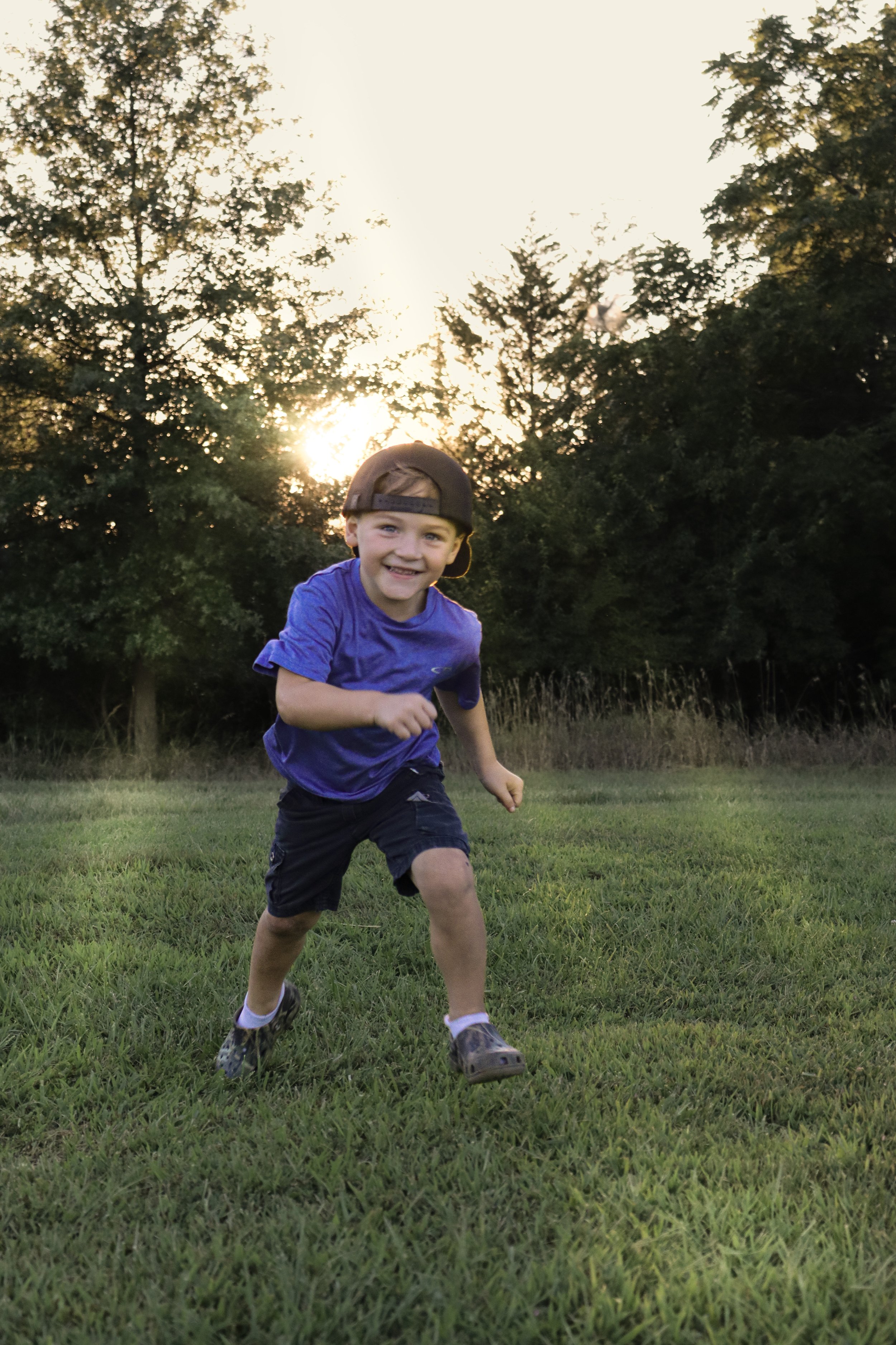 A young boy running and smiling on a grassy field during sunset, wearing a purple shirt, dark shorts, a cap worn backwards, and running shoes.