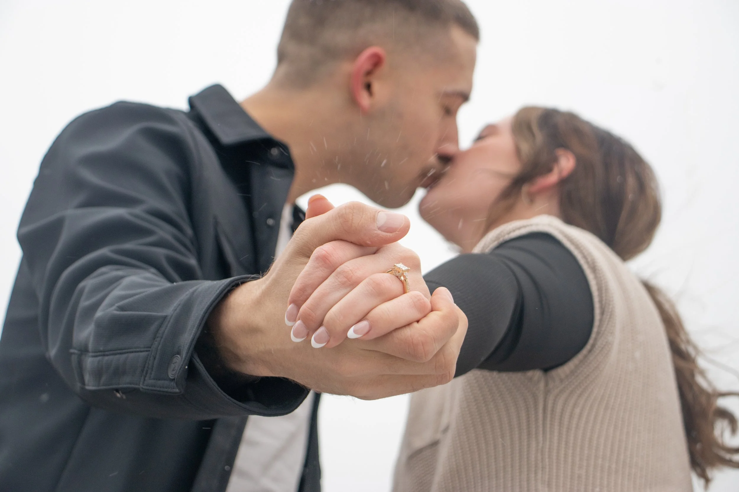 A couple sharing a kiss, holding hands with a gold ring prominently displayed, with a plain white background.