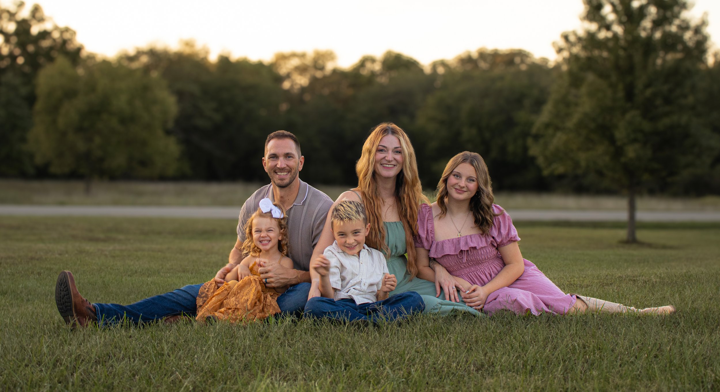 A family sitting on the grass in a park with trees and a sunset in the background, smiling at the camera.