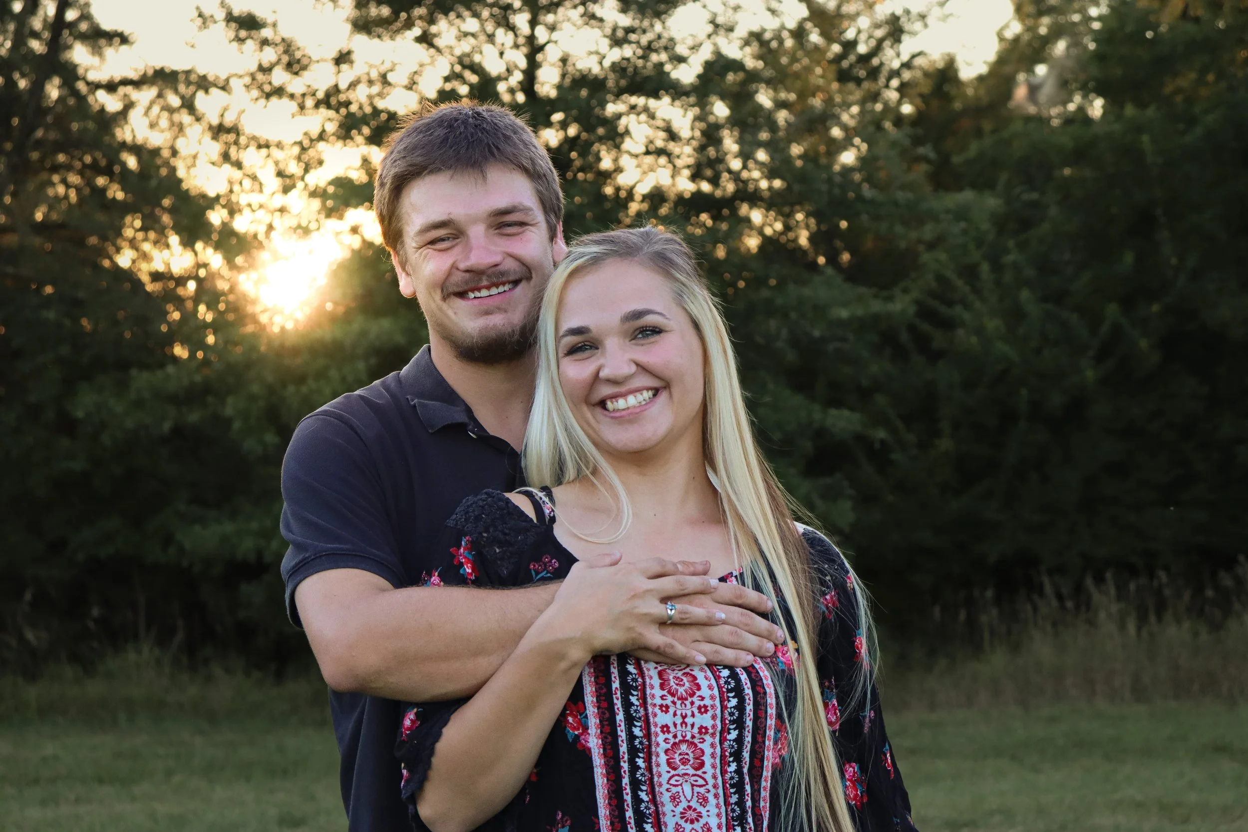 A smiling couple standing together outdoors on a grassy field during sunset, with trees in the background.