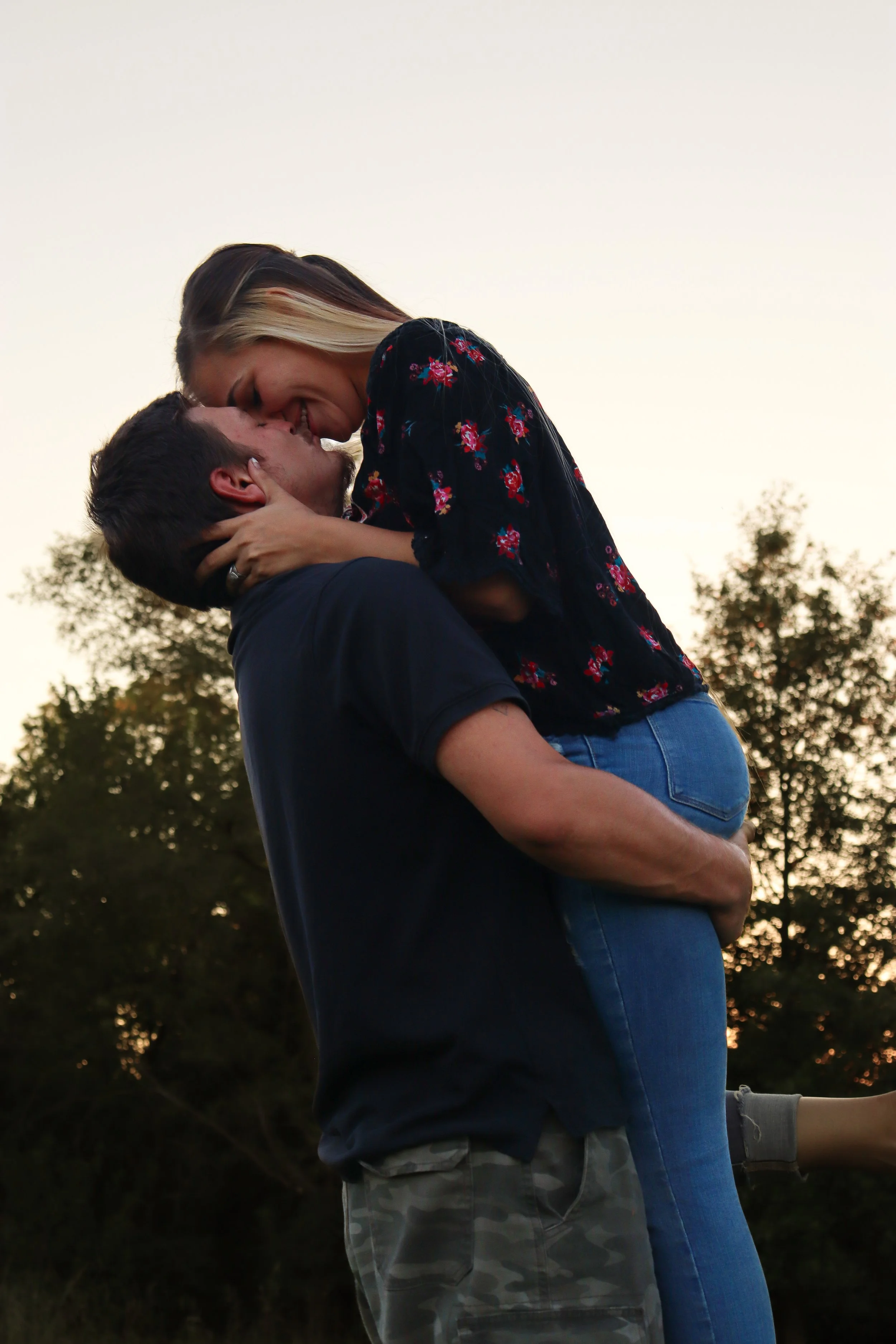 A man lifting a woman in an outdoor setting during sunset, with trees in the background, both smiling and close to each other.