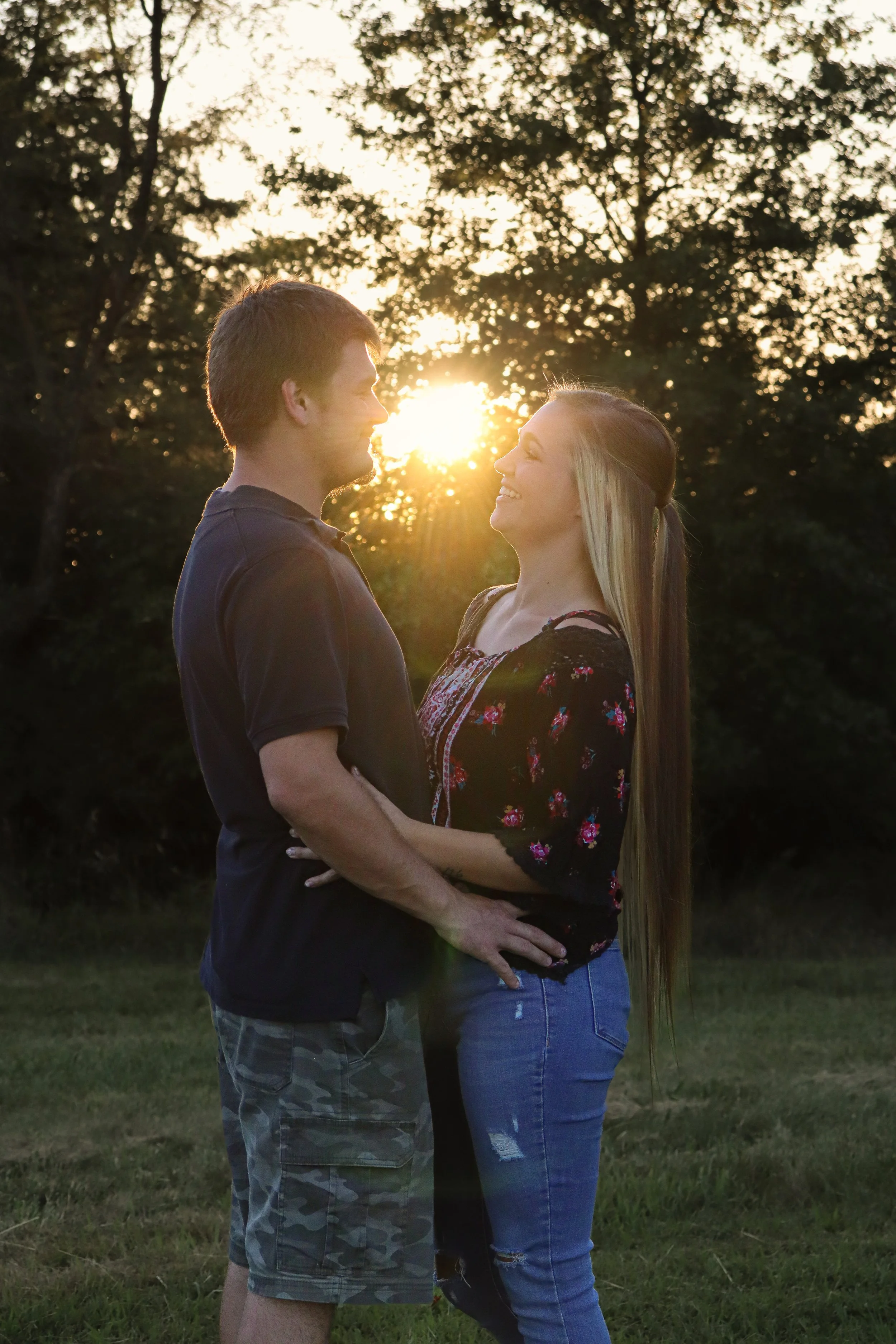 A couple standing outdoors in a park at sunset, smiling and looking at each other, with trees in the background.