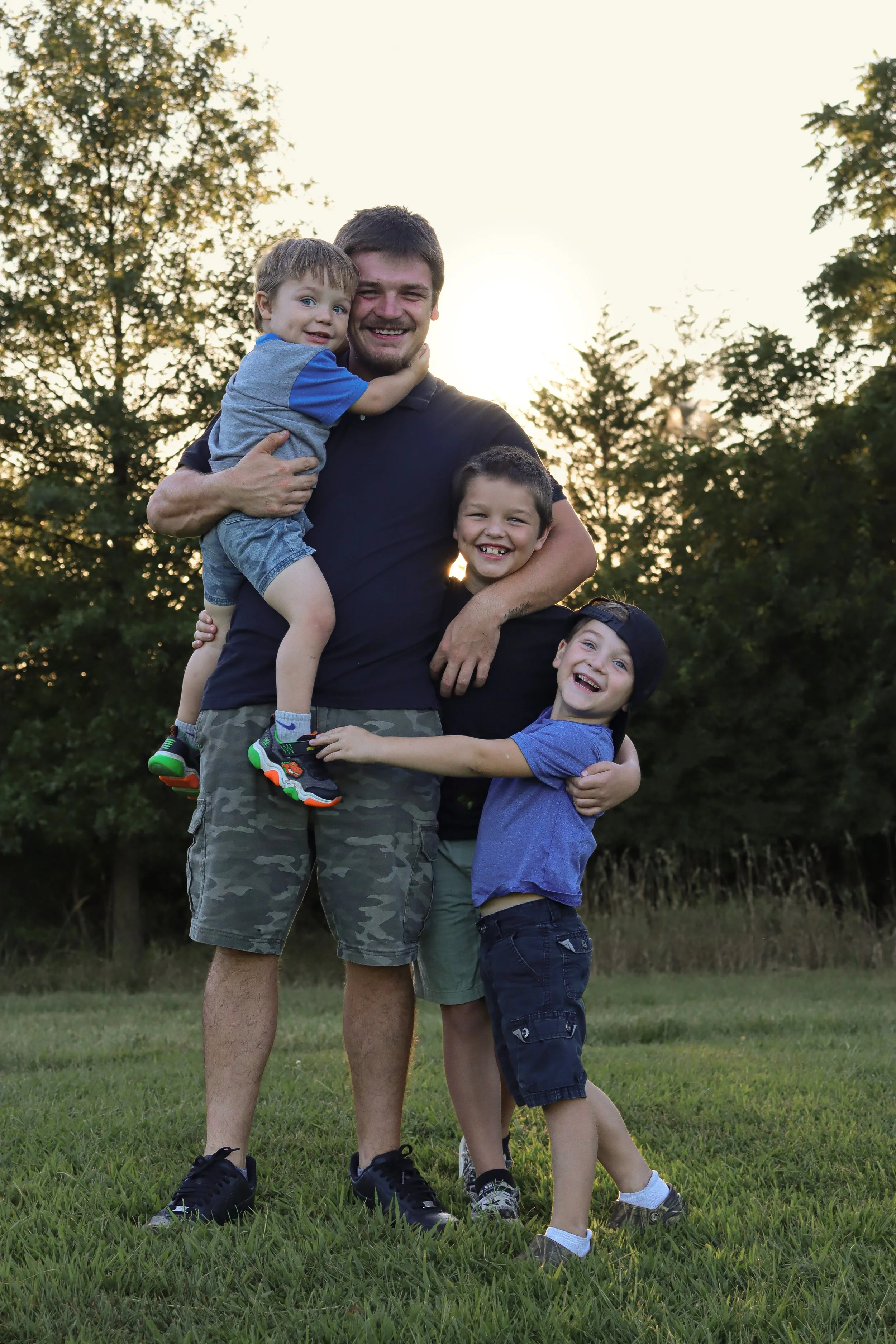 A man and three boys hugging outdoors at sunset in a grassy field with trees in the background.