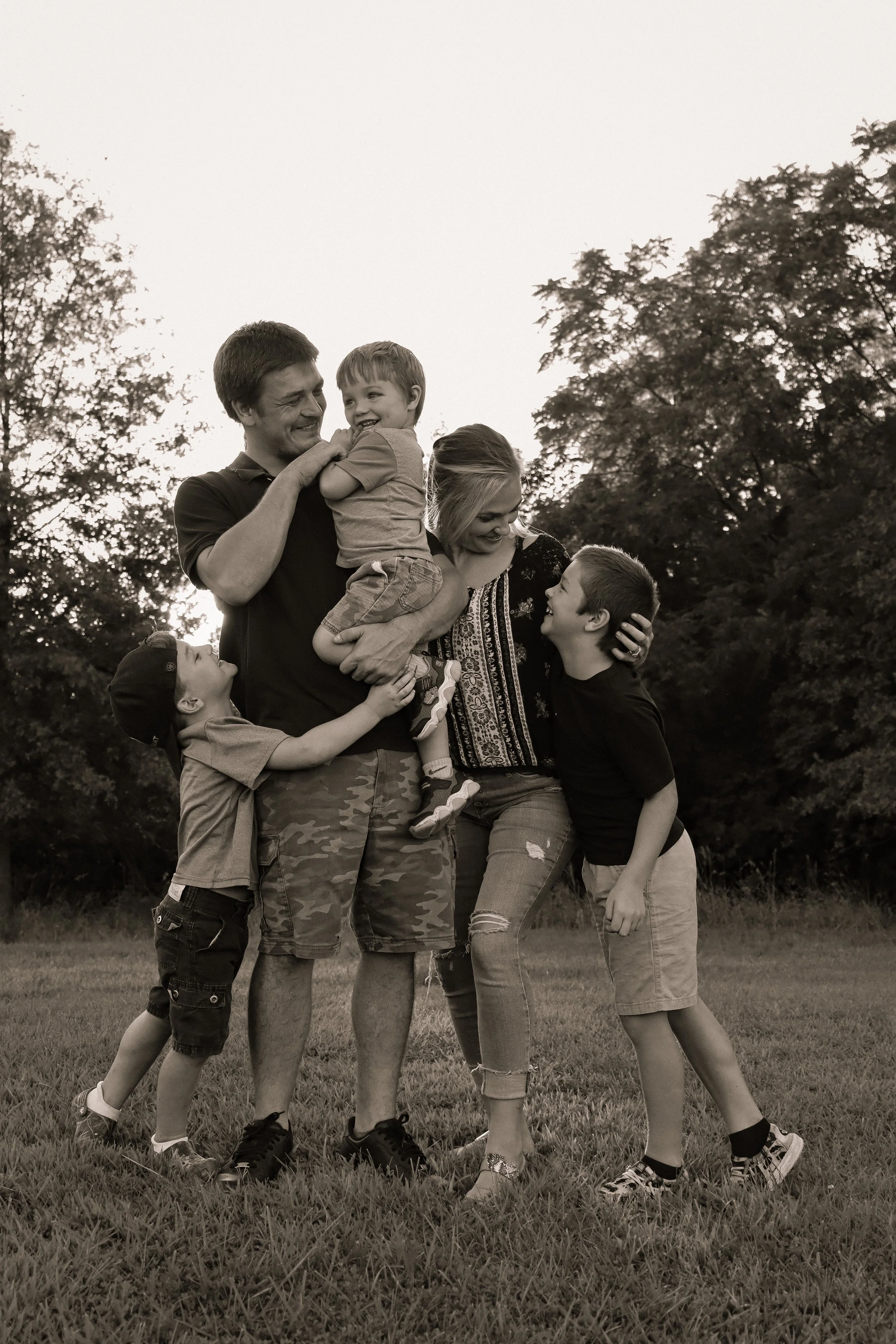A family of six, including two adults and four children, enjoying a playful moment outdoors on a grassy field with trees in the background.
