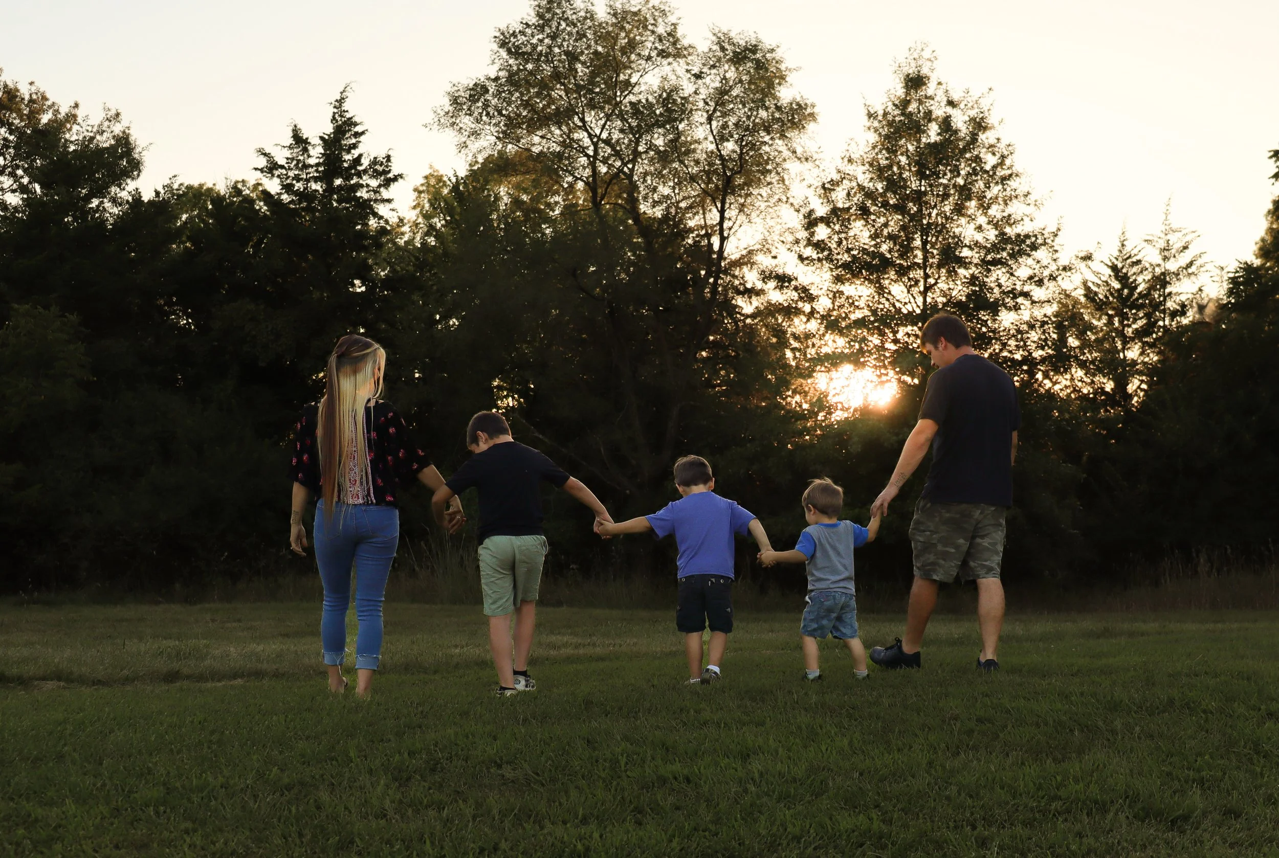 A family, including two adults and four children, holding hands and walking across a grassy field at sunset with trees in the background.