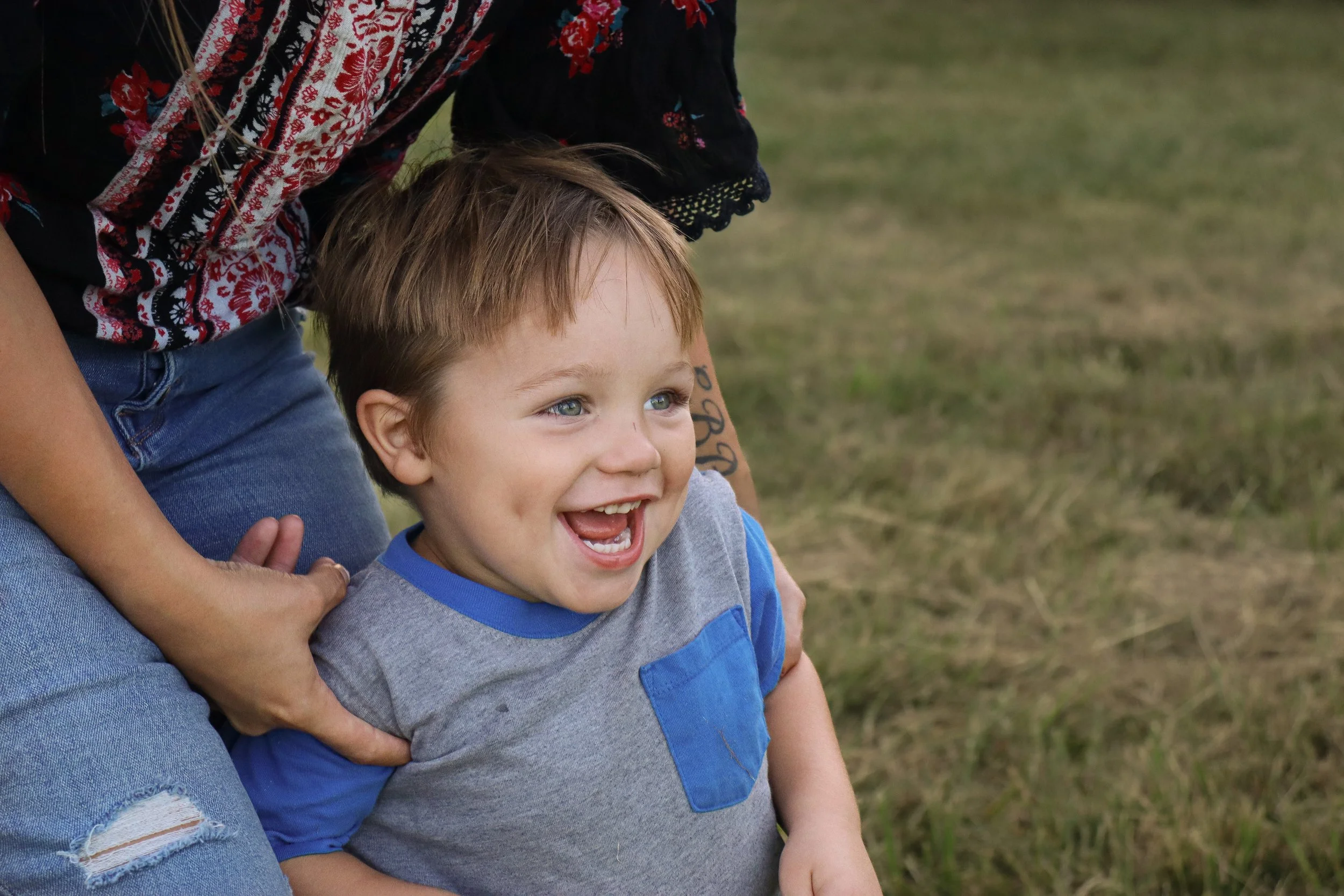 A young boy with blue eyes and brown hair smiling and playing outdoors, with a person in a floral shirt and jeans holding him from behind.
