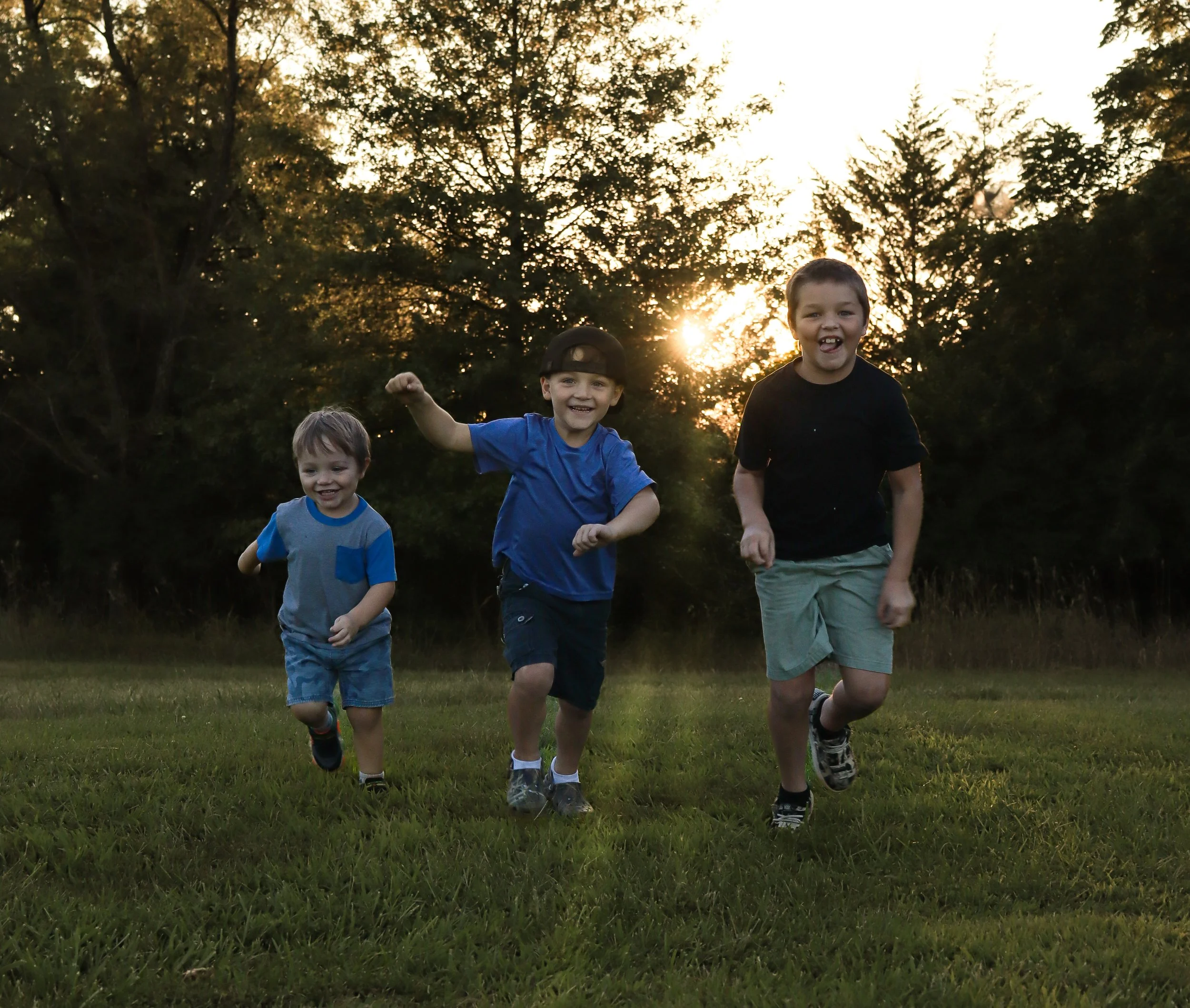 Three boys running and smiling in a grassy field during sunset with trees in the background.