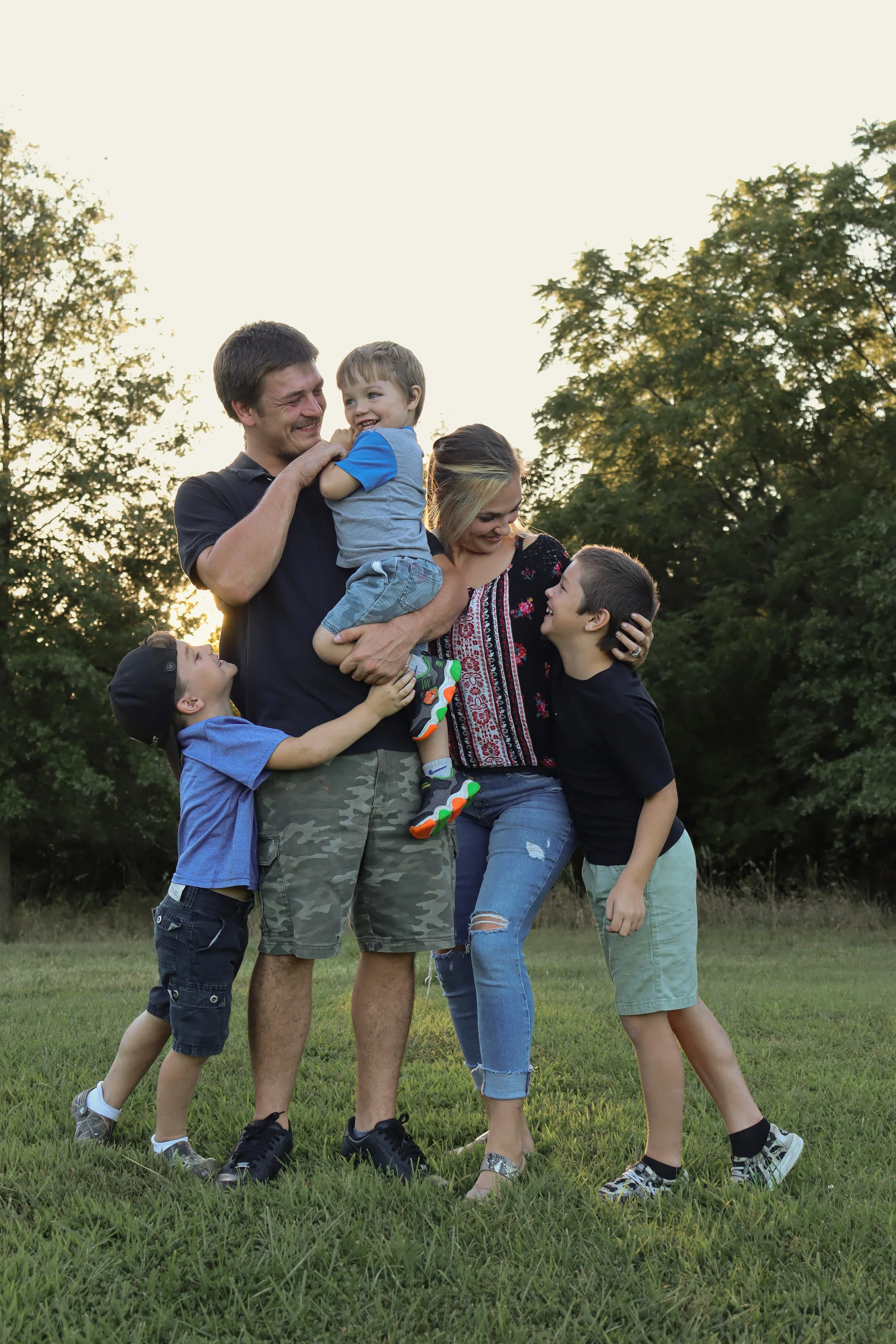A family of five, including parents and three children, standing outdoors on grass, smiling and playing together during sunset.