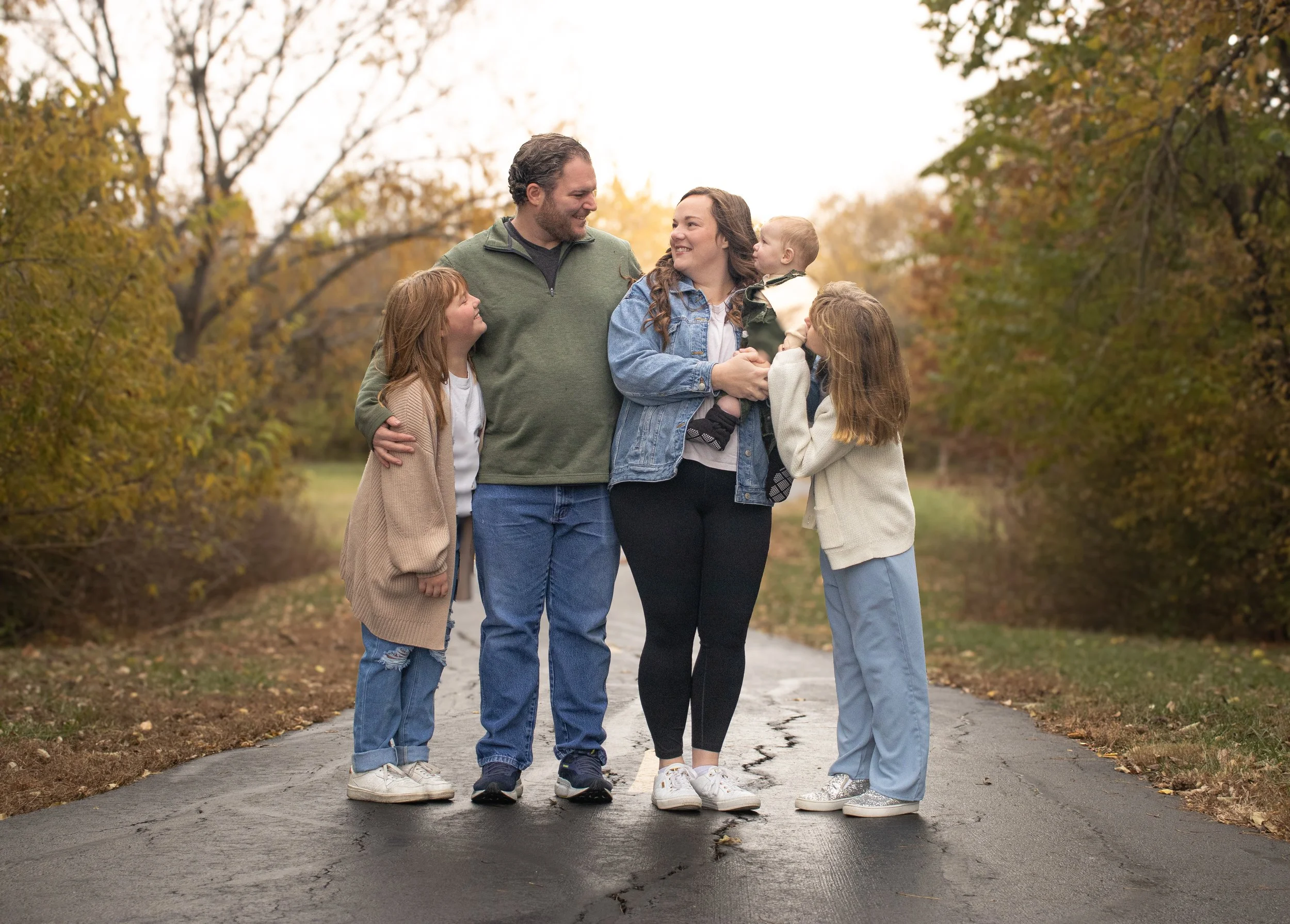 Family of six standing on a paved path in a park during autumn with colorful fall trees, smiling and looking at each other.