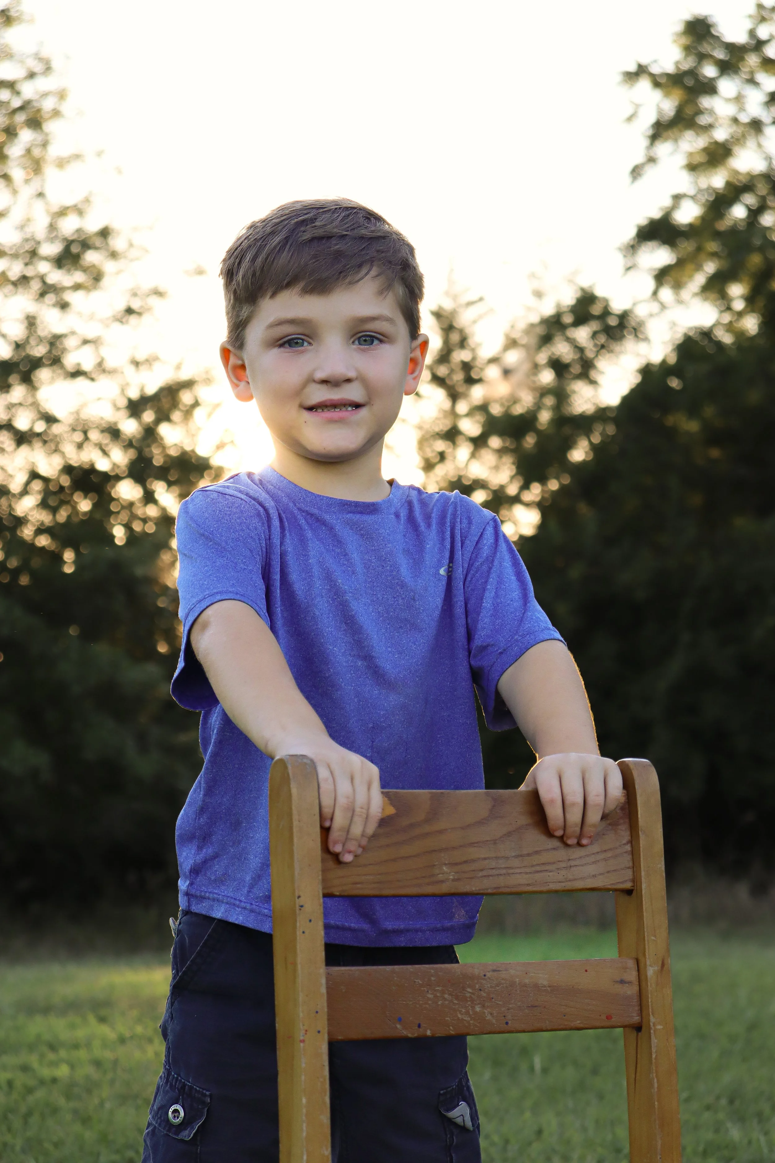 A young boy in a purple t-shirt standing outdoors at sunset, holding onto a wooden chair, with trees in the background.
