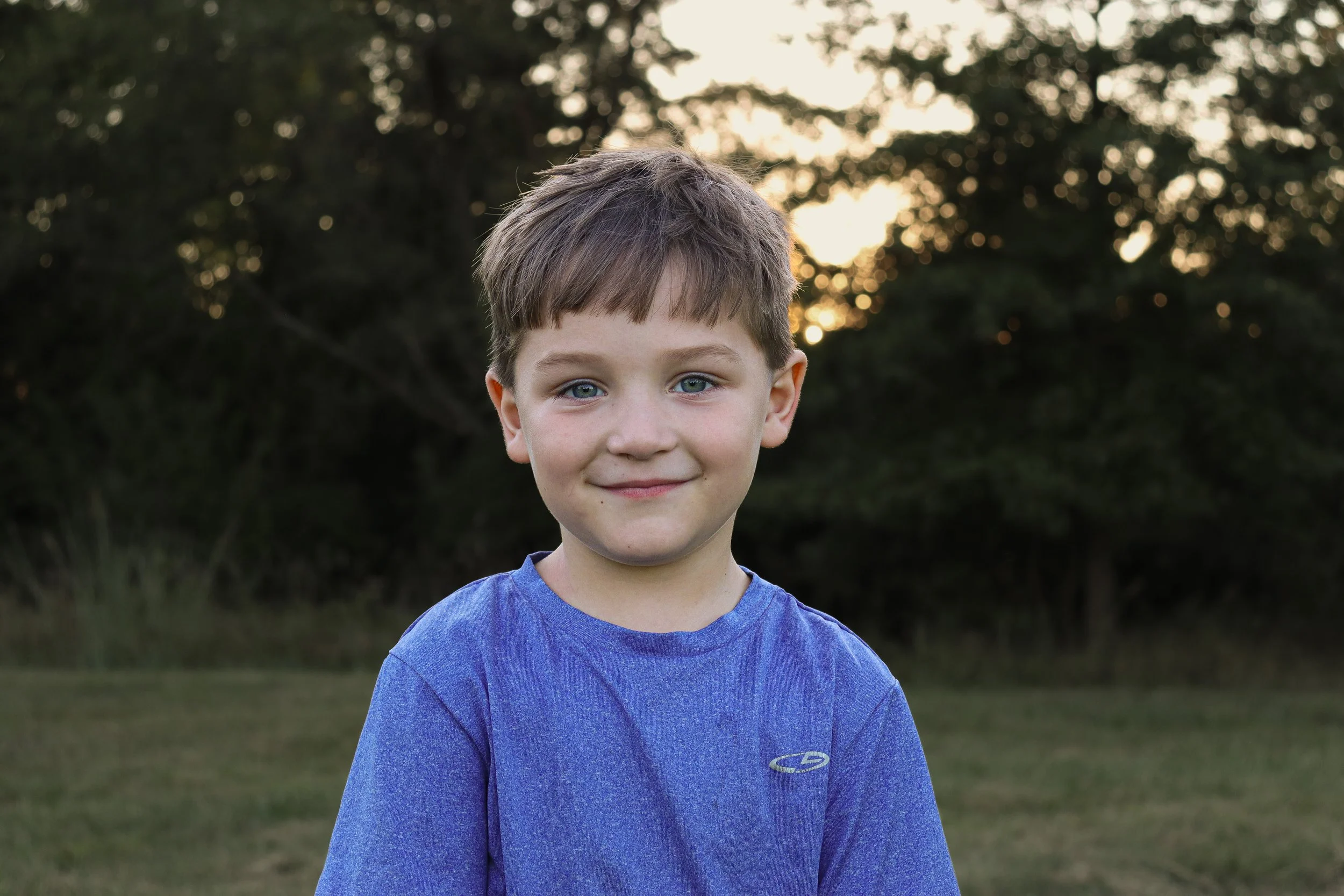 A young boy with blue eyes and light brown hair smiling outdoors during sunset, wearing a blue shirt, with trees in the background.