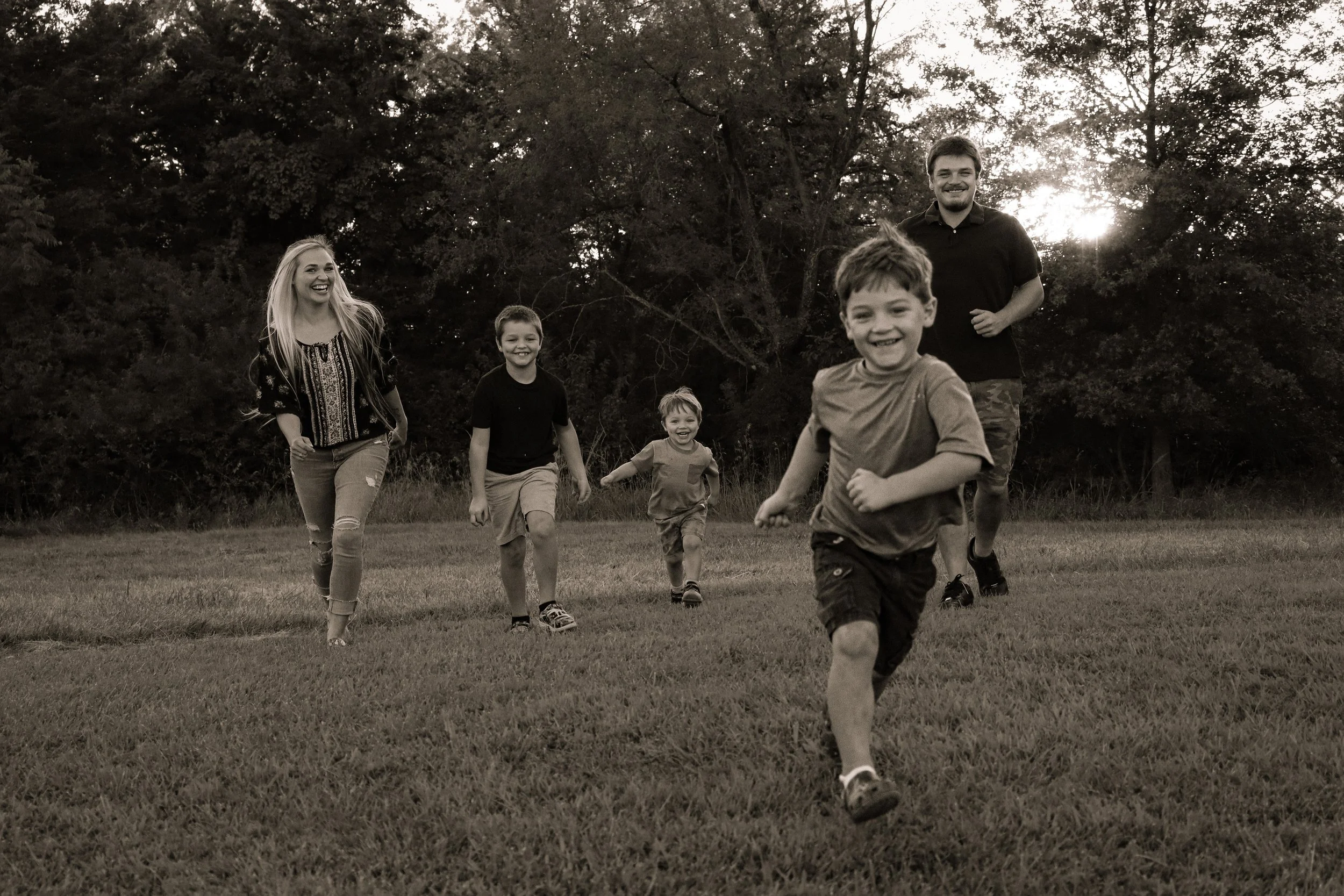 A group of six people, including children and two adults, running and smiling in a grassy field at sunset with trees in the background.