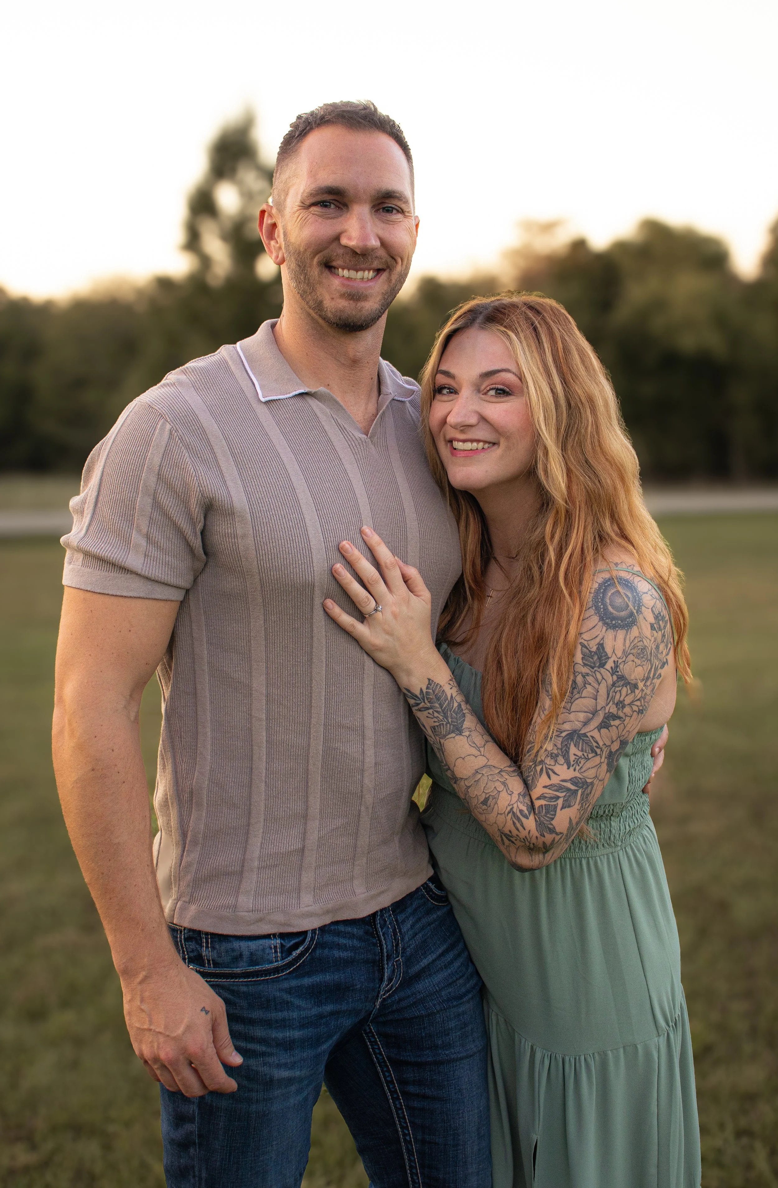 A smiling couple standing outdoors on a grassy field at sunset, with trees in the background. The woman has long red hair, tattoos on her arm, and is wearing a green dress, while the man has short dark hair and is wearing a beige polo shirt and jeans. She is showing her left hand with a ring.