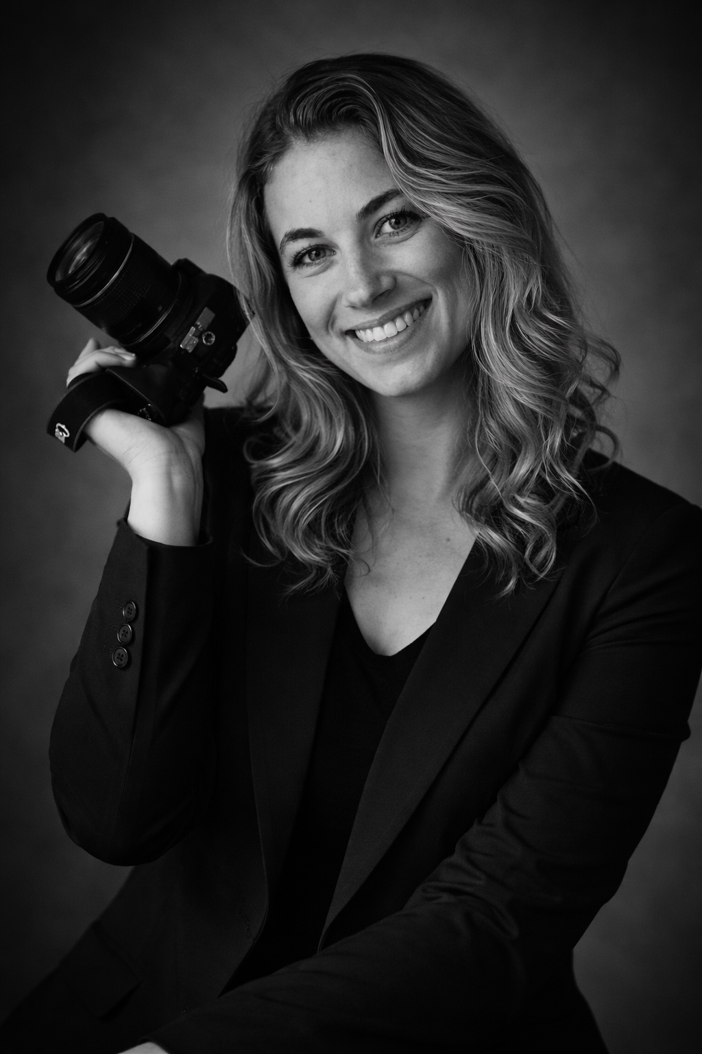 A portrait of a smiling woman with wavy hair wearing a black blazer, holding a camera on her shoulder in black and white.