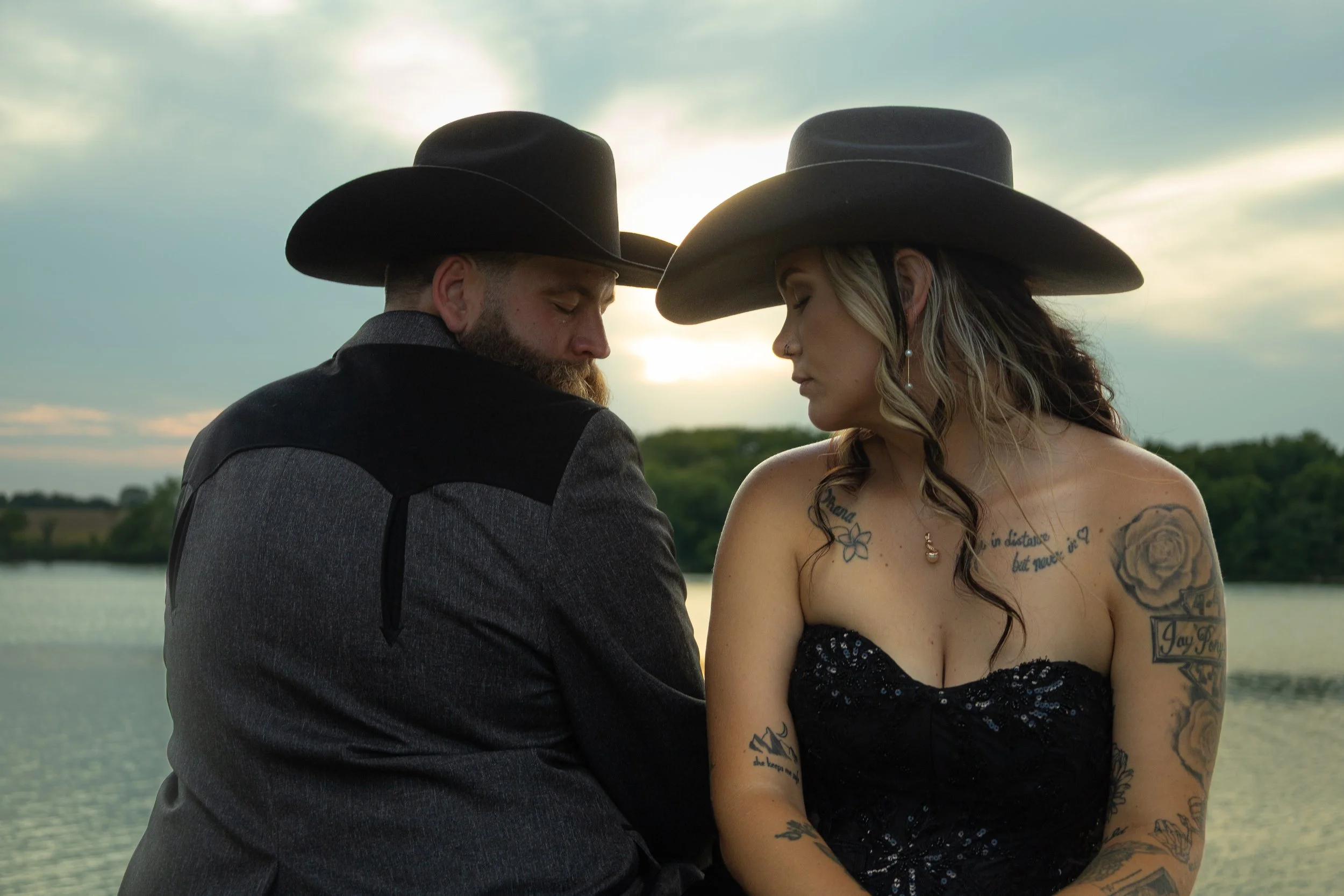 A man and woman wearing cowboy hats, sitting close together by a river at sunset, with their heads bowed and eyes closed.
