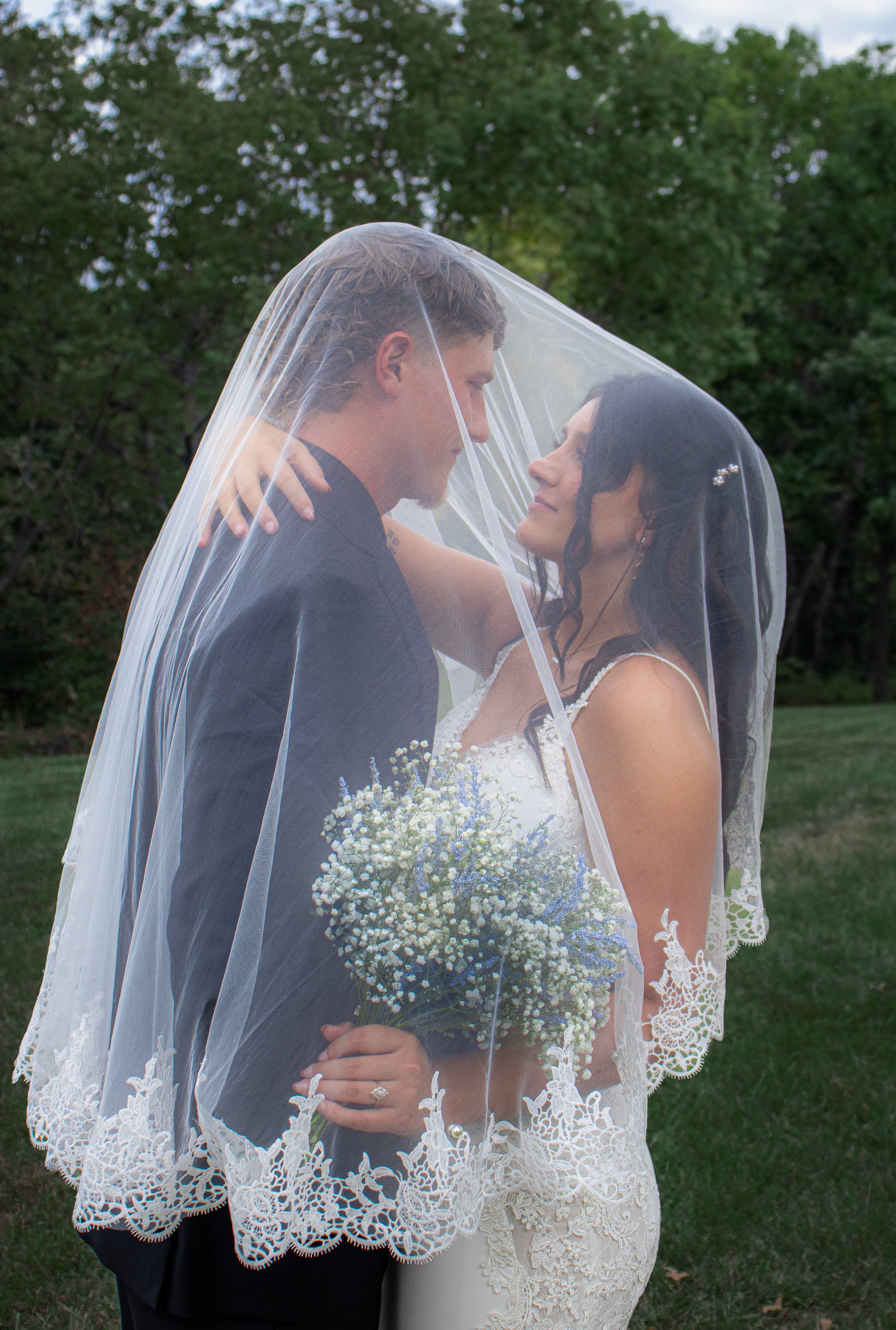 A bride and groom under a wedding veil in an outdoor setting, with the bride holding a bouquet of white and blue flowers and the groom wearing a black suit.