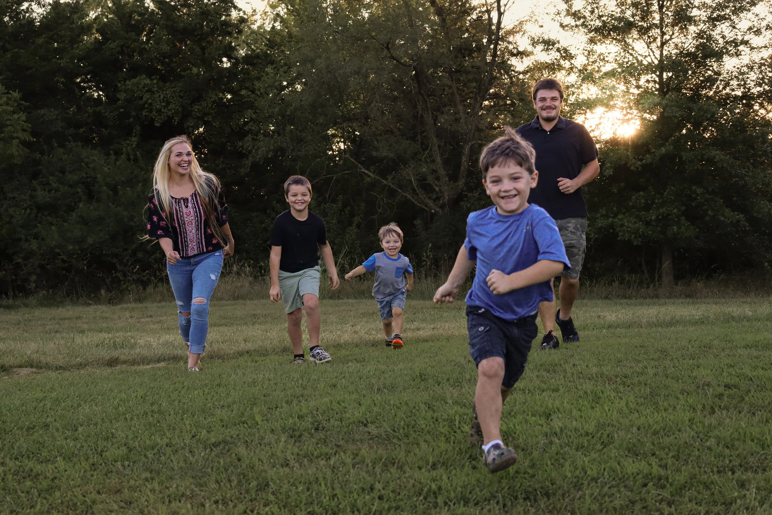 A family of six running and playing together outdoors on a grassy field during sunset, with trees in the background.
