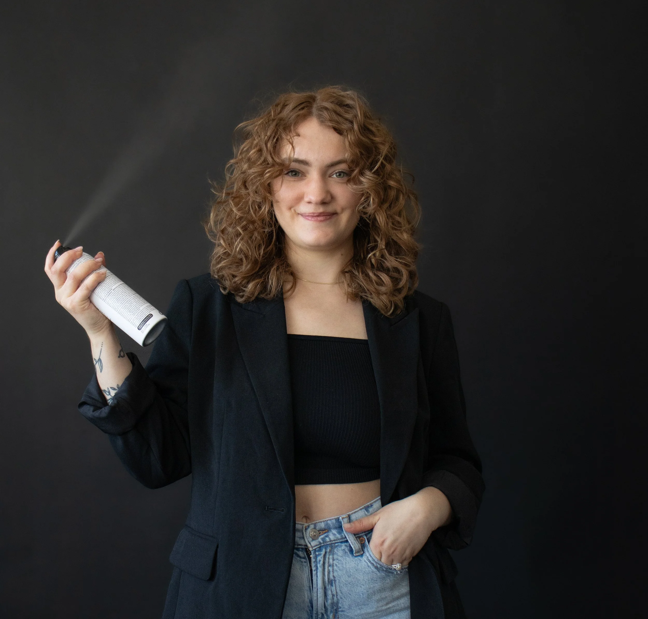 Young woman with curly red hair holding a spray can behind her head against a dark background, smiling at the camera.