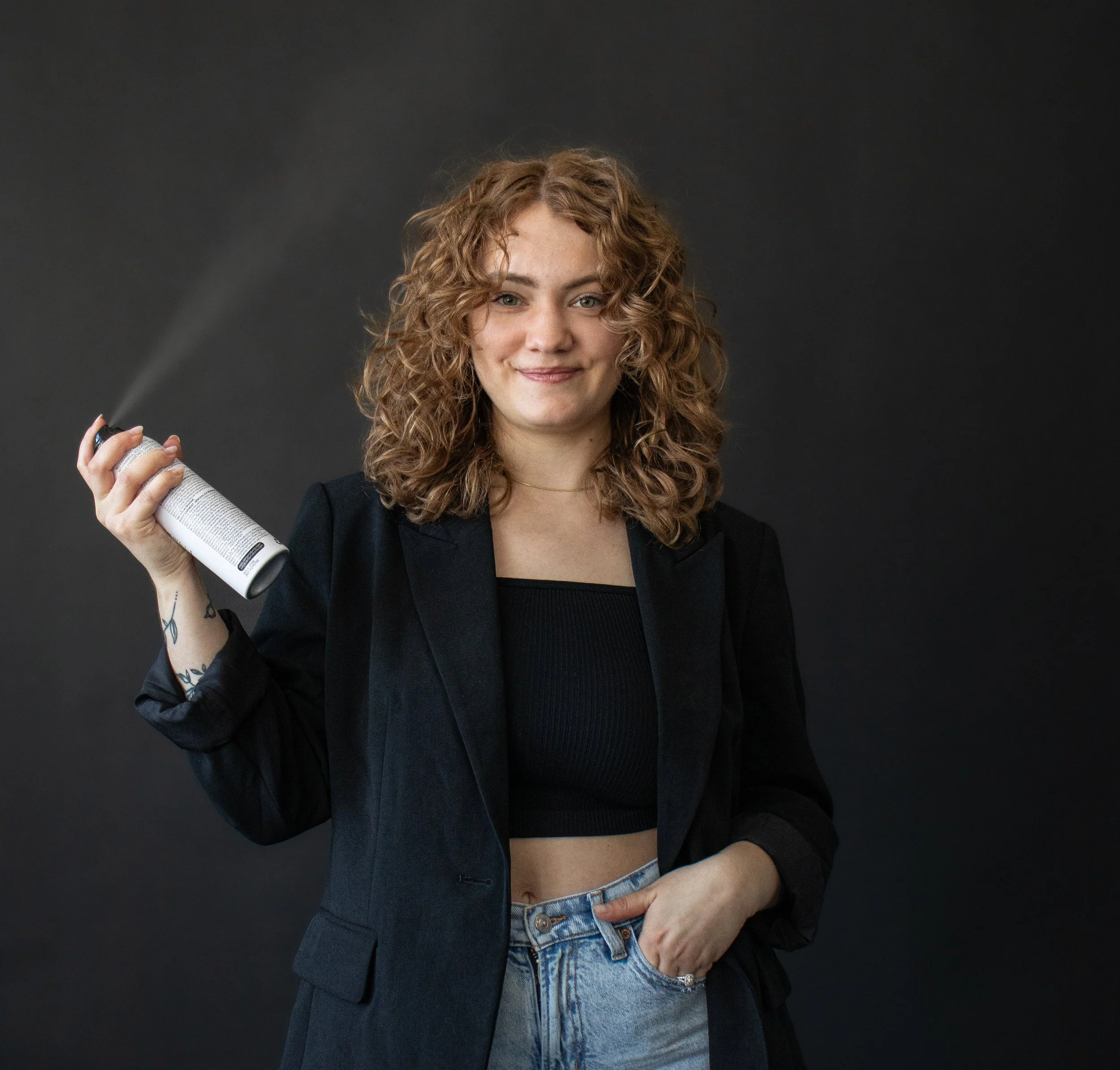 A young woman with curly red hair wearing a black blazer and black crop top, holding a spray can in her right hand, with a faint mist coming out, standing against a dark background.