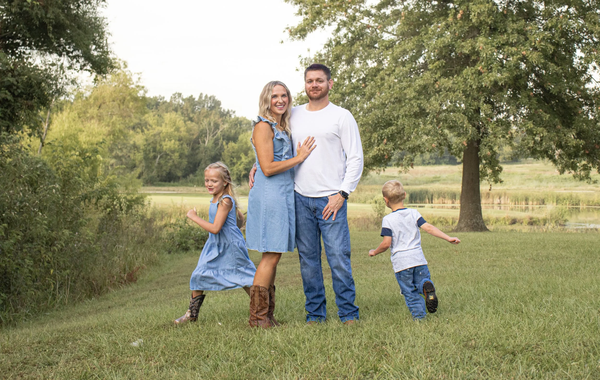 A family of four standing on a grassy field with trees and a pond in the background. The mother and father are embracing and smiling at the camera, while their young daughter and son are playing nearby, with the daughter in a blue dress and cowboy boots, and the son in a gray shirt and jeans running away.