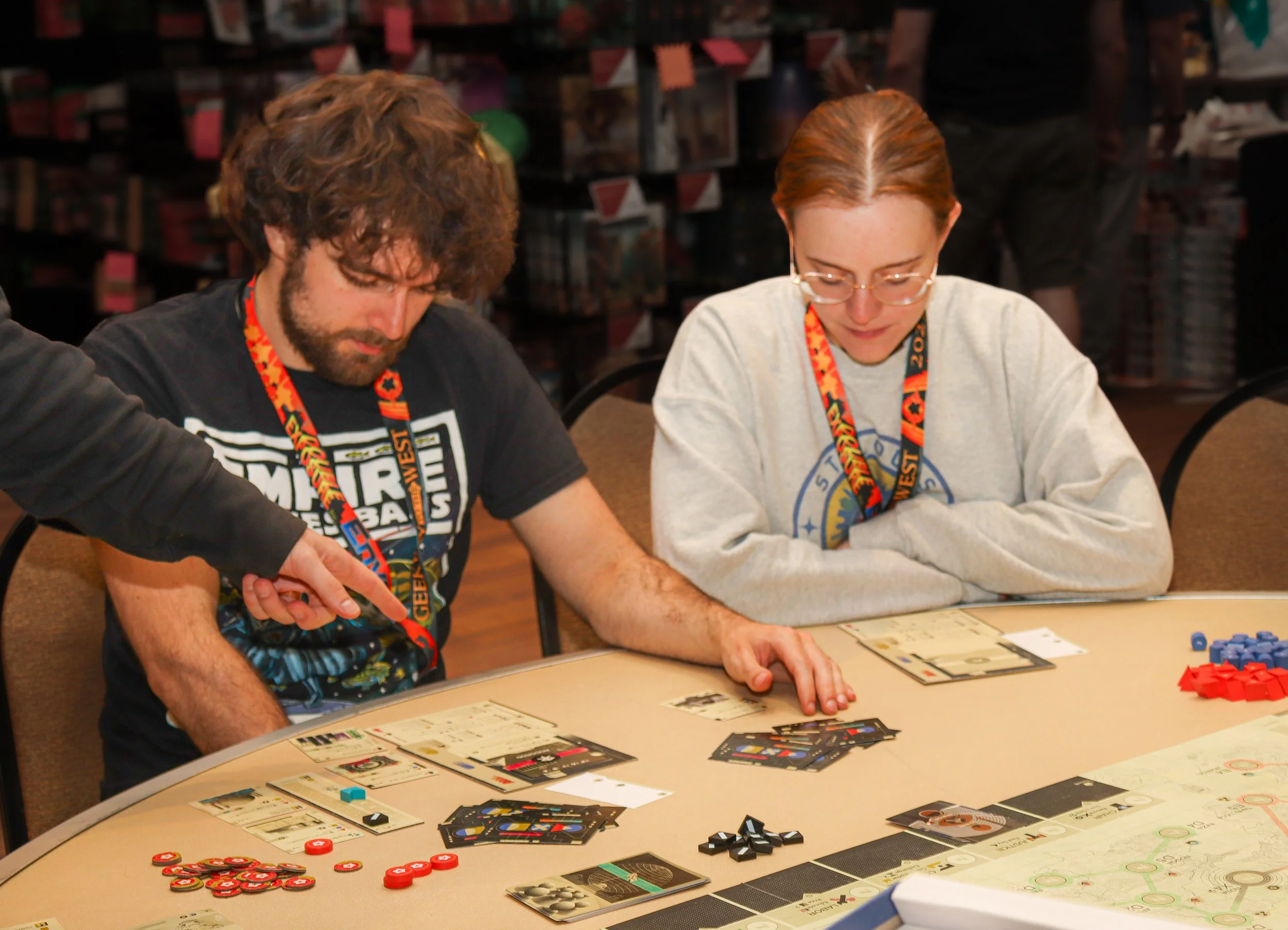 Two people sit at a table playing a board game, with game pieces, cards, and tokens visible. One person appears focused, and the other is looking down at the game. A person is pointing at the game.