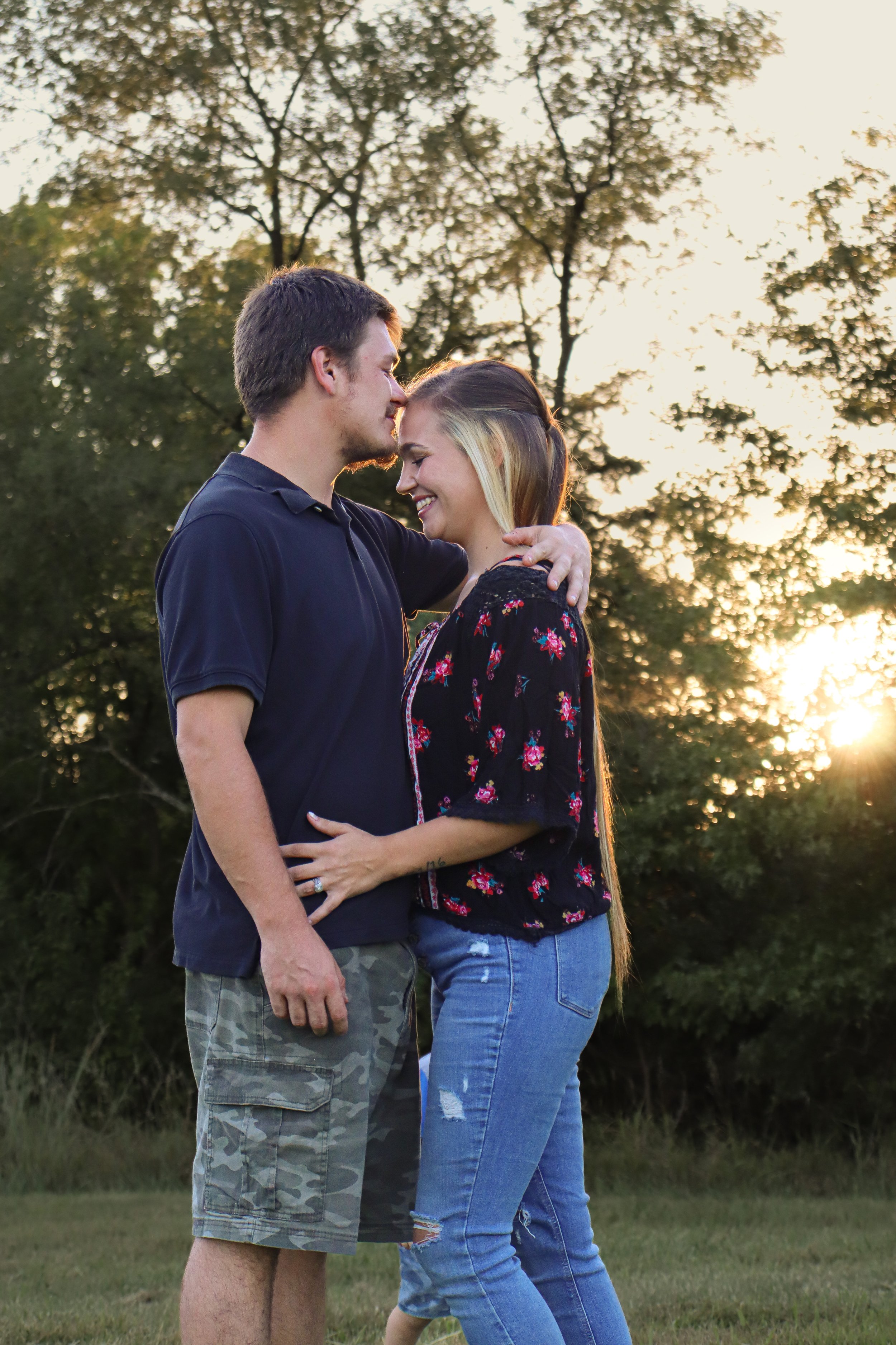 A young couple standing closely together outdoors during sunset, smiling and embracing with trees in the background.