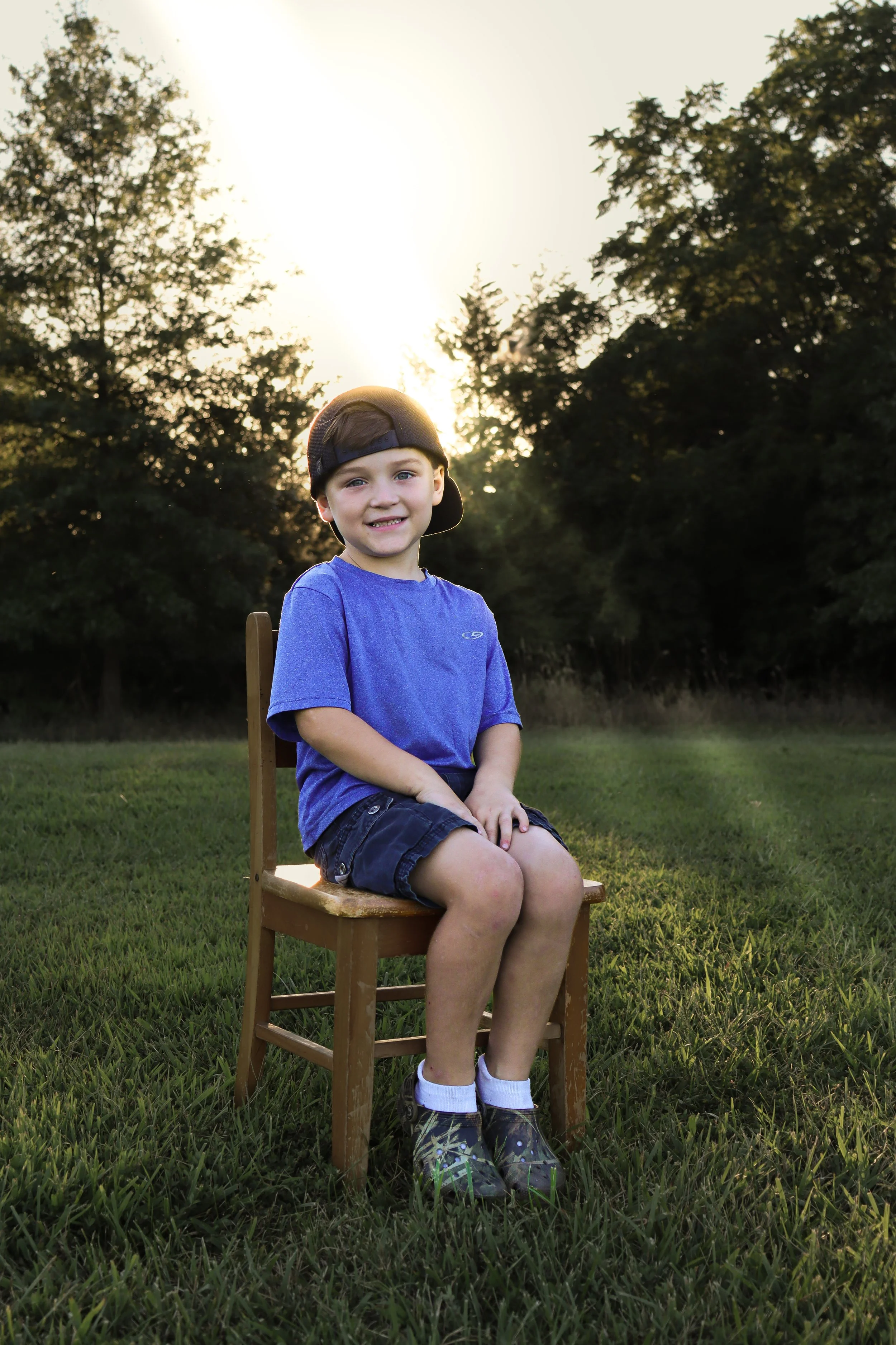 A young boy sitting on a wooden chair outdoors during sunset, wearing a blue t-shirt, dark shorts, and sneakers, with trees in the background.