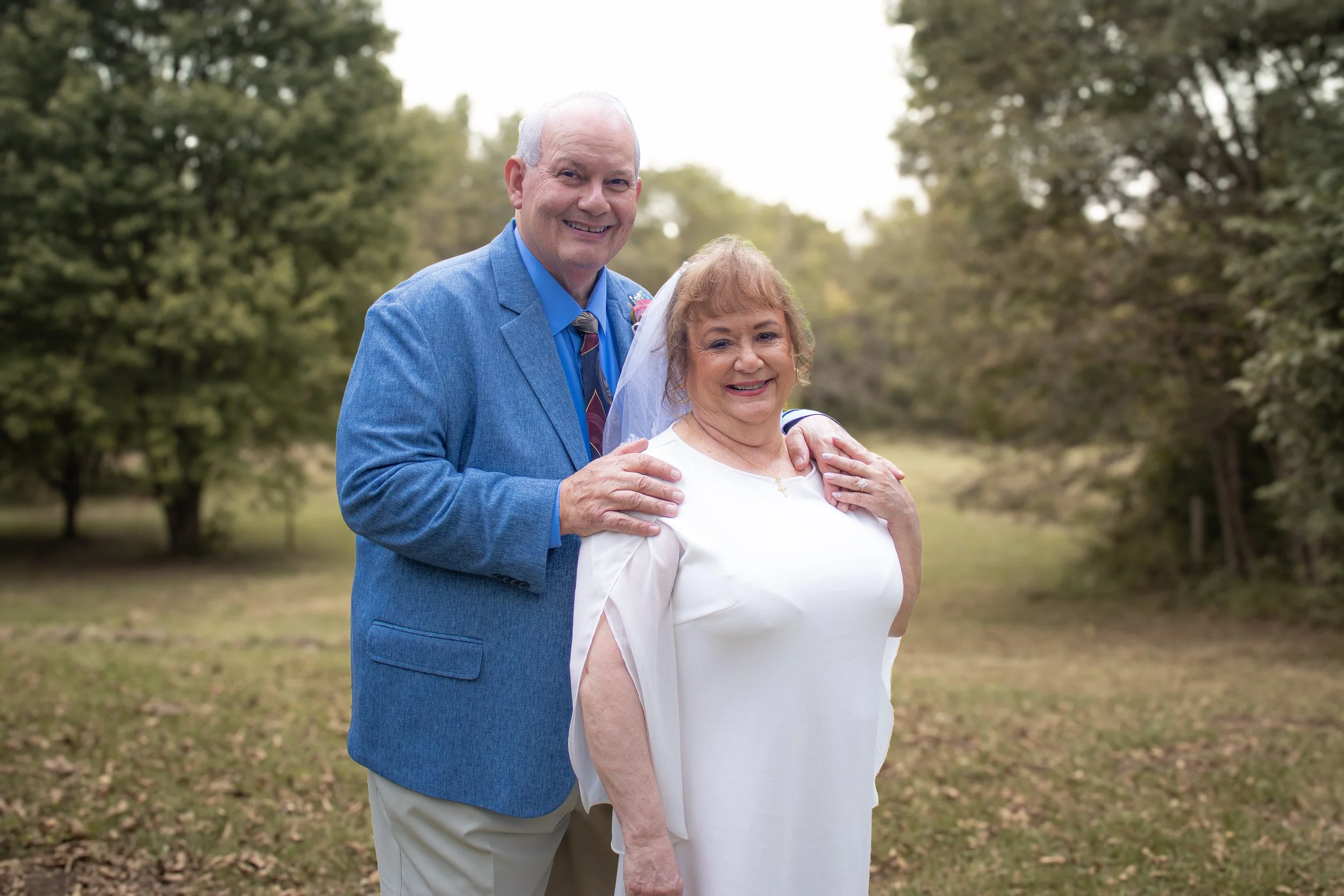 An elderly couple standing outdoors in a park, smiling at the camera, with trees and a grassy path in the background. The man has white hair and is wearing a blue blazer, blue shirt, and patterned tie. The woman has light brown hair and is dressed in a white dress with a veil, suggesting a wedding or special occasion.