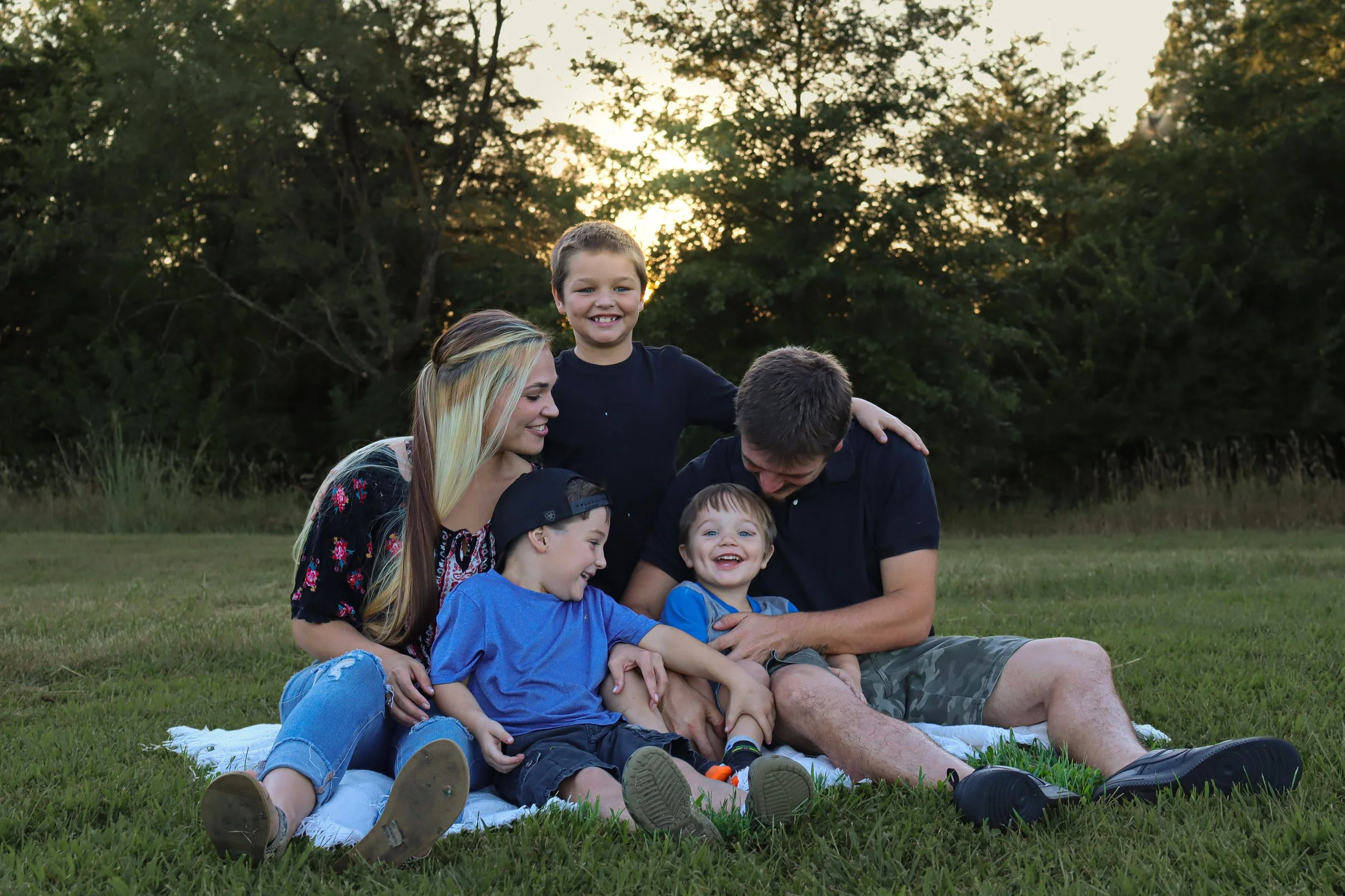 A family of six sitting on a blanket in a grassy field at sunset, happily playing and smiling together.