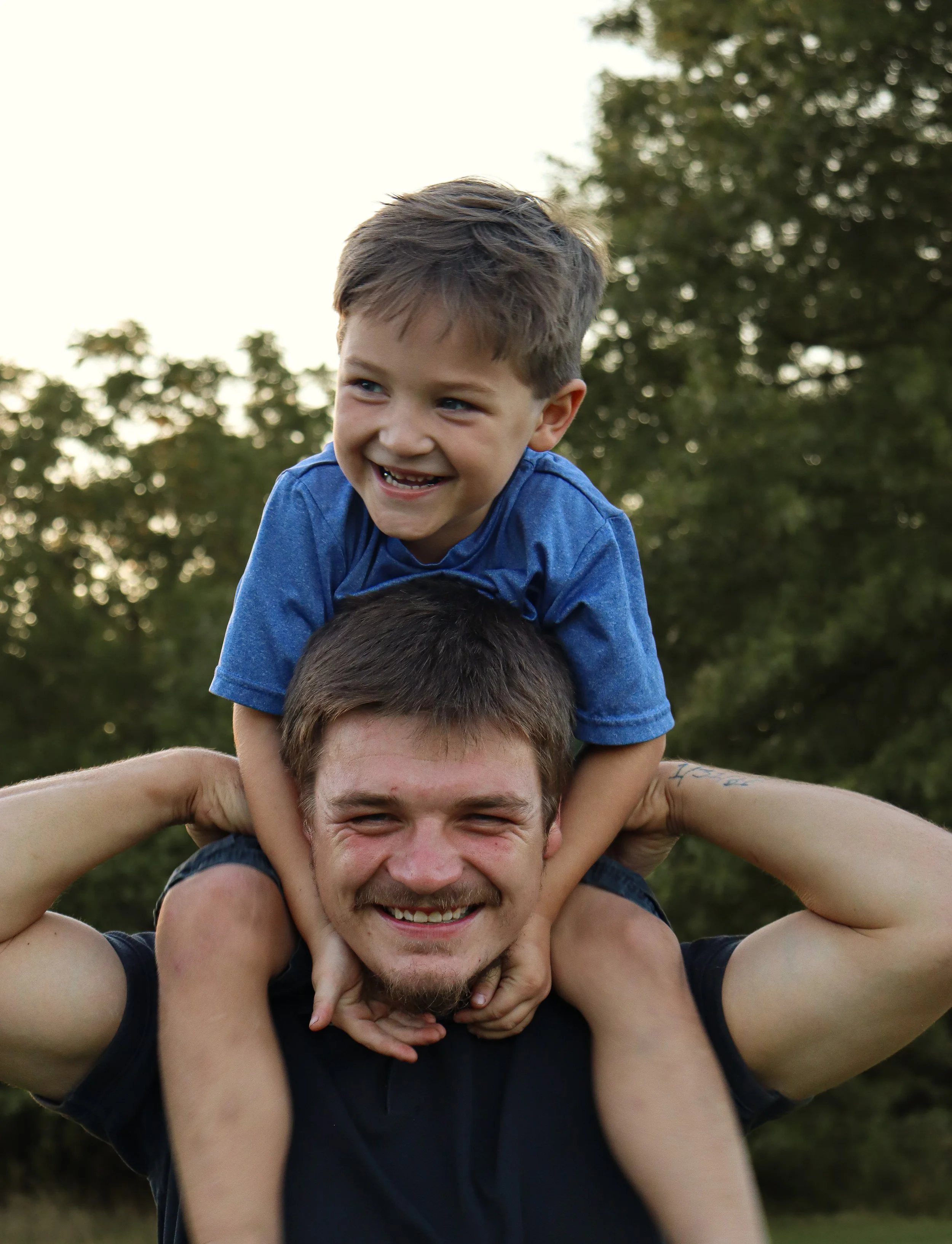 A man and a young boy smiling, with the boy sitting on the man's shoulders outdoors near trees.