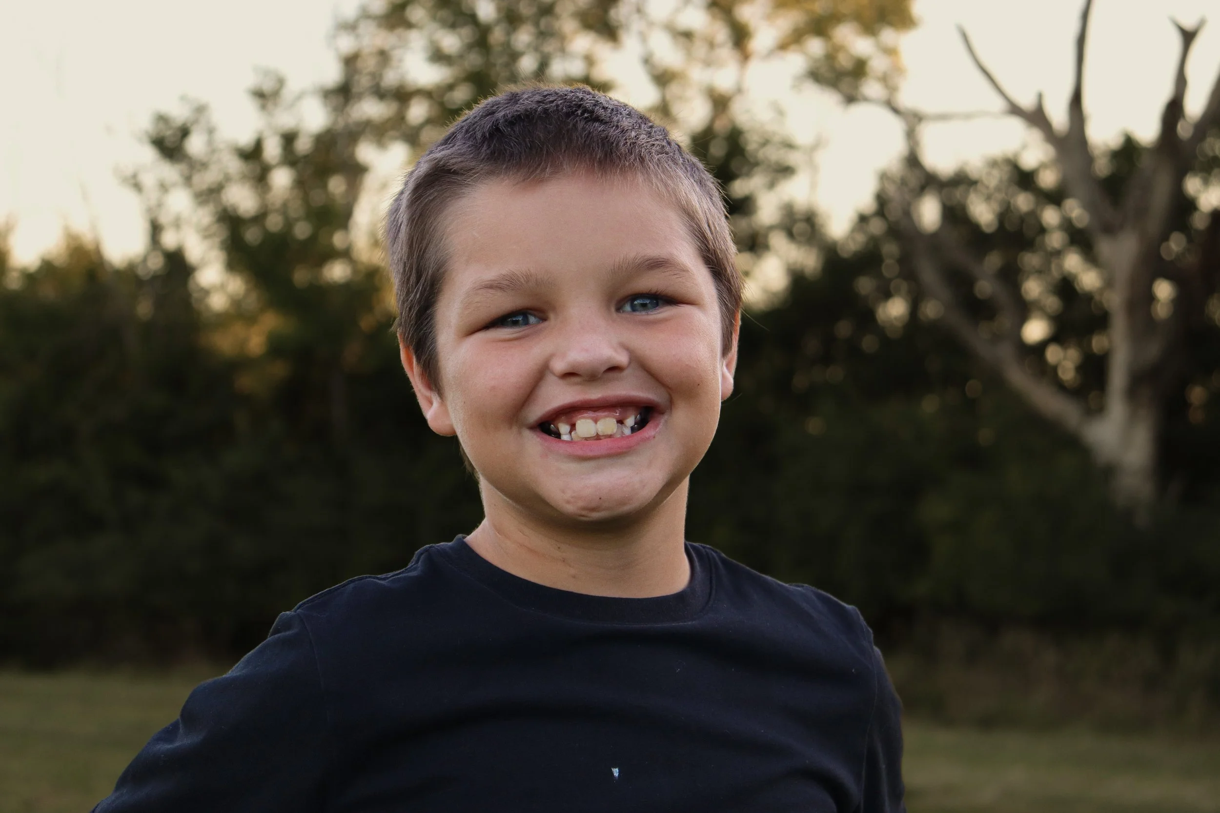 A young boy with short brown hair, blue eyes, and missing front teeth smiling outdoors with trees and a partly cloudy sky in the background.