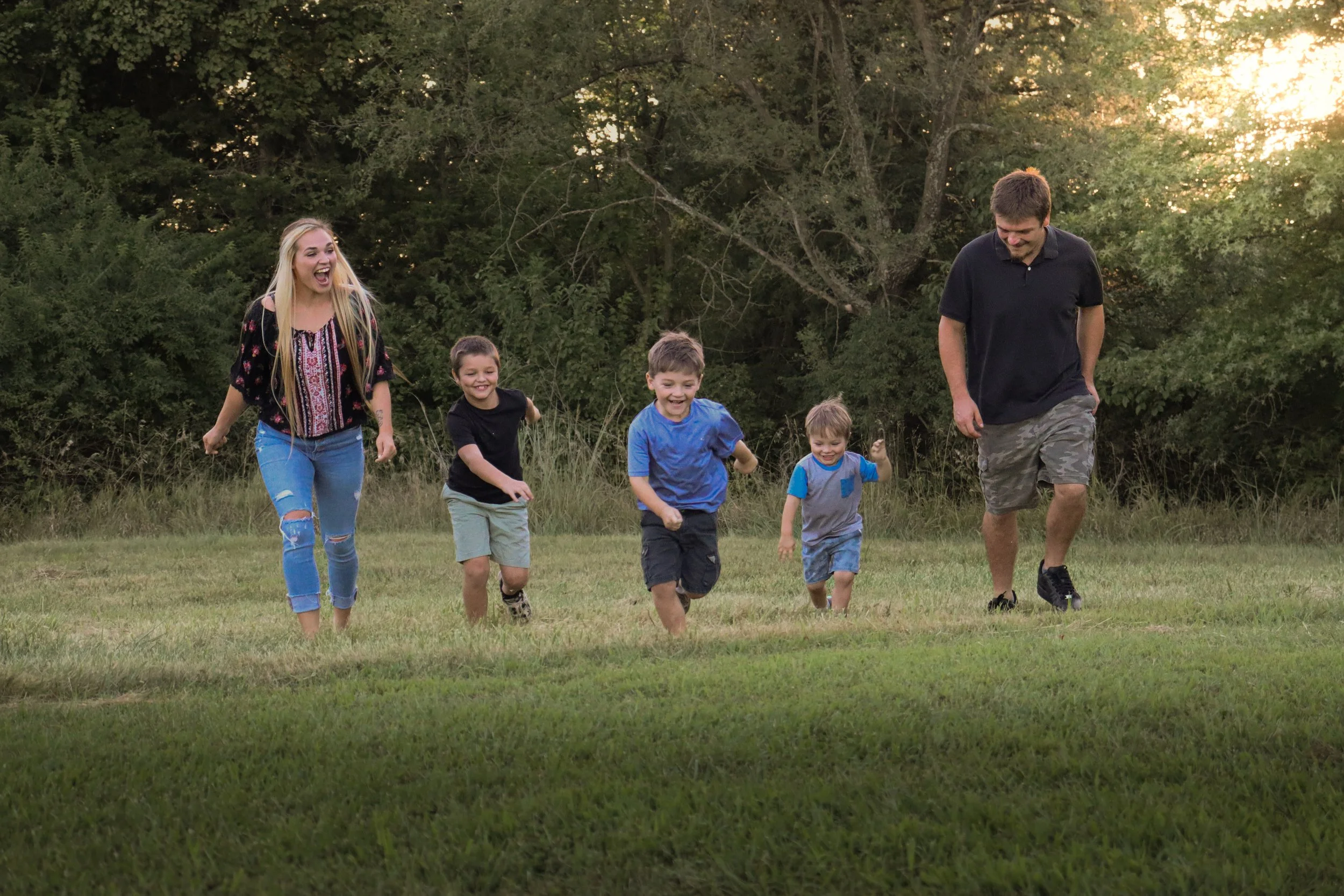 A family of six, including two adults and four children, runs and laughs on a grassy field during sunset with trees in the background.