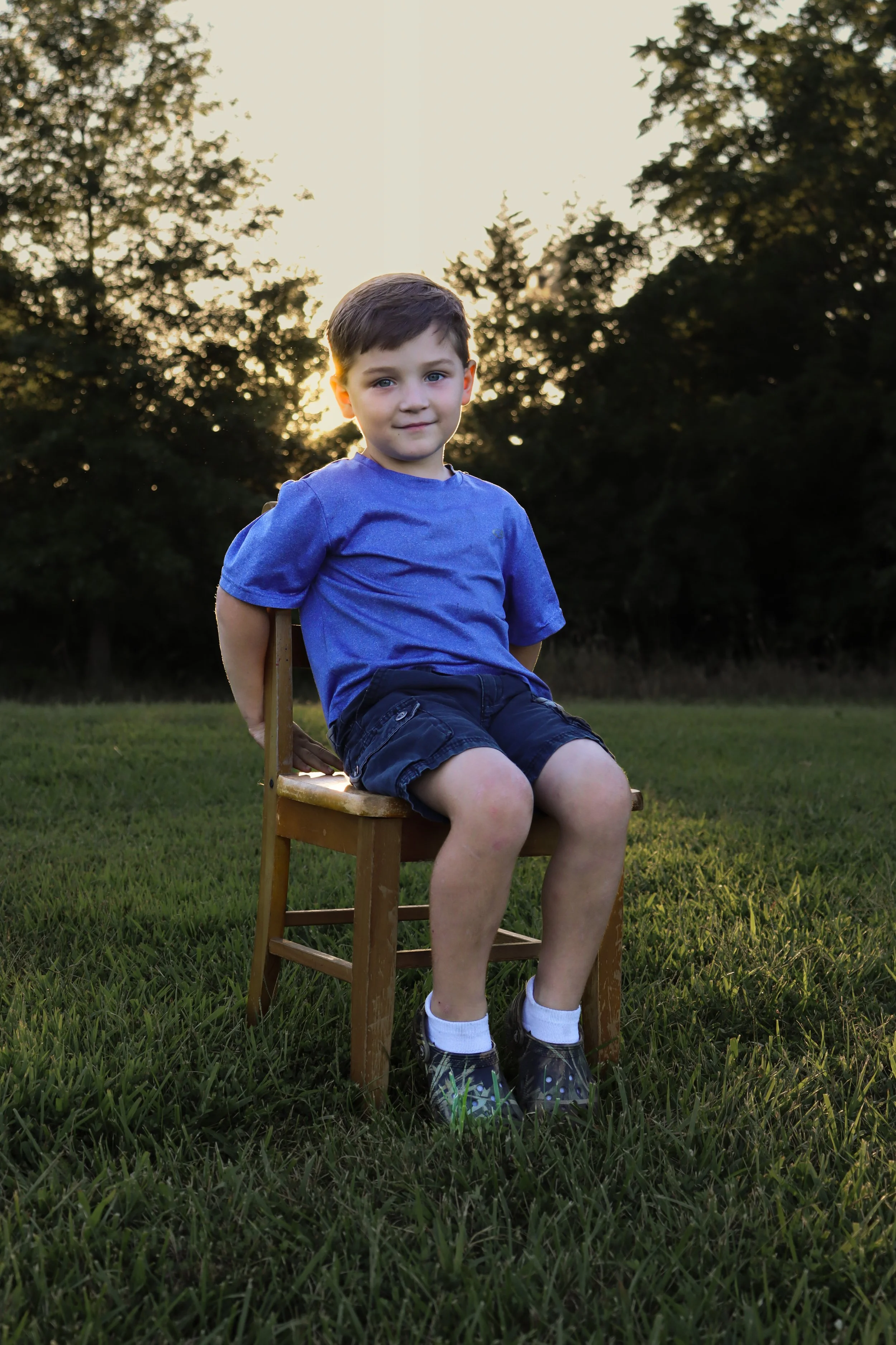 A young boy sitting on a wooden chair on a grassy field during sunset, wearing a blue t-shirt, black shorts, white socks, and black shoes.