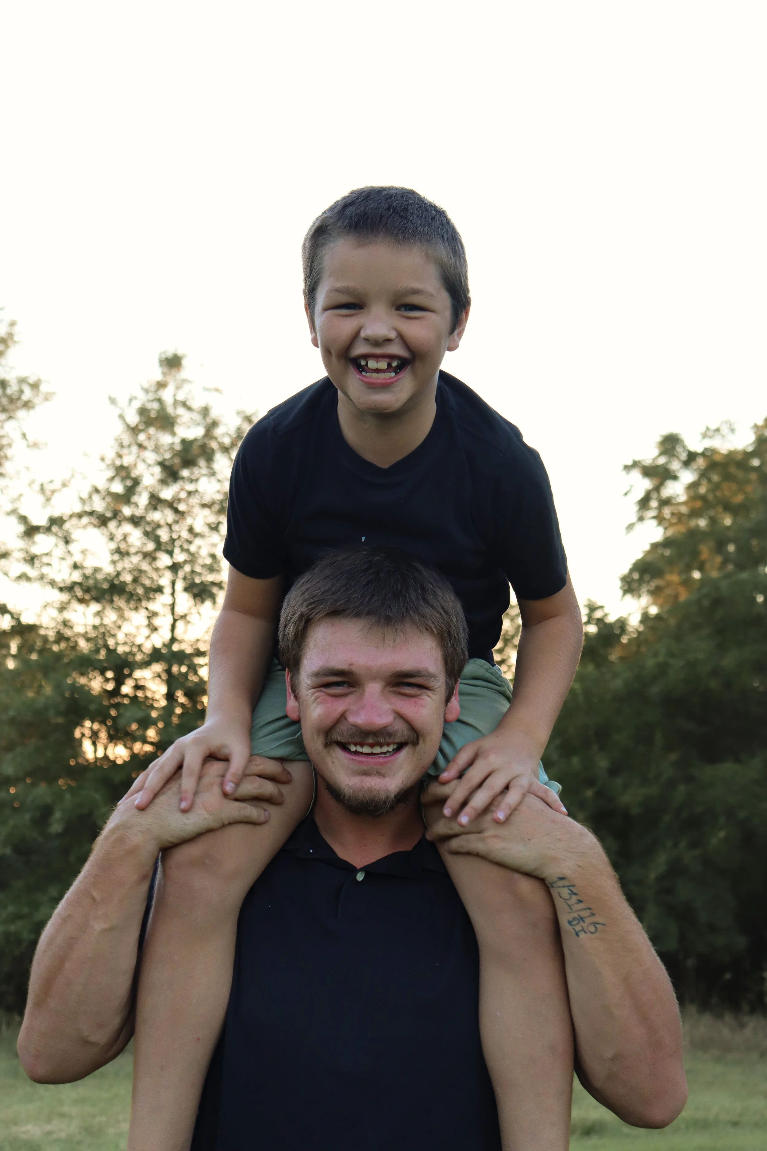 A man and a young boy smiling, the boy sitting on the man's shoulders outdoors during sunset.