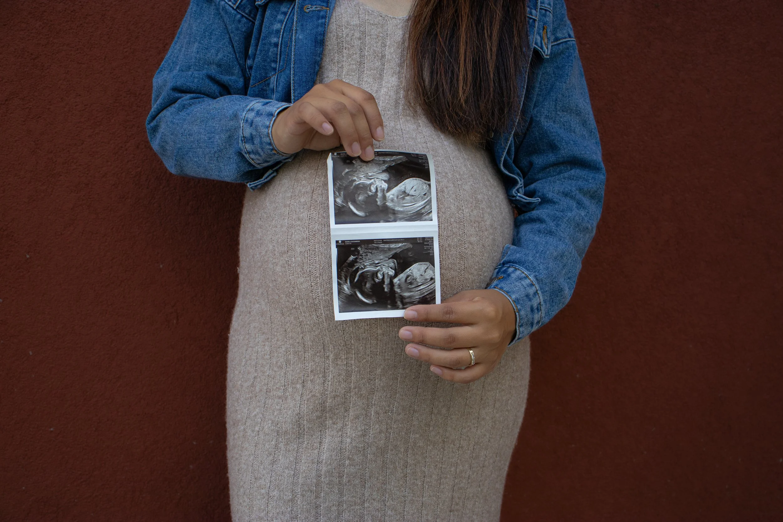 A pregnant woman holding two ultrasound images in front of her abdomen, standing against a red wall.