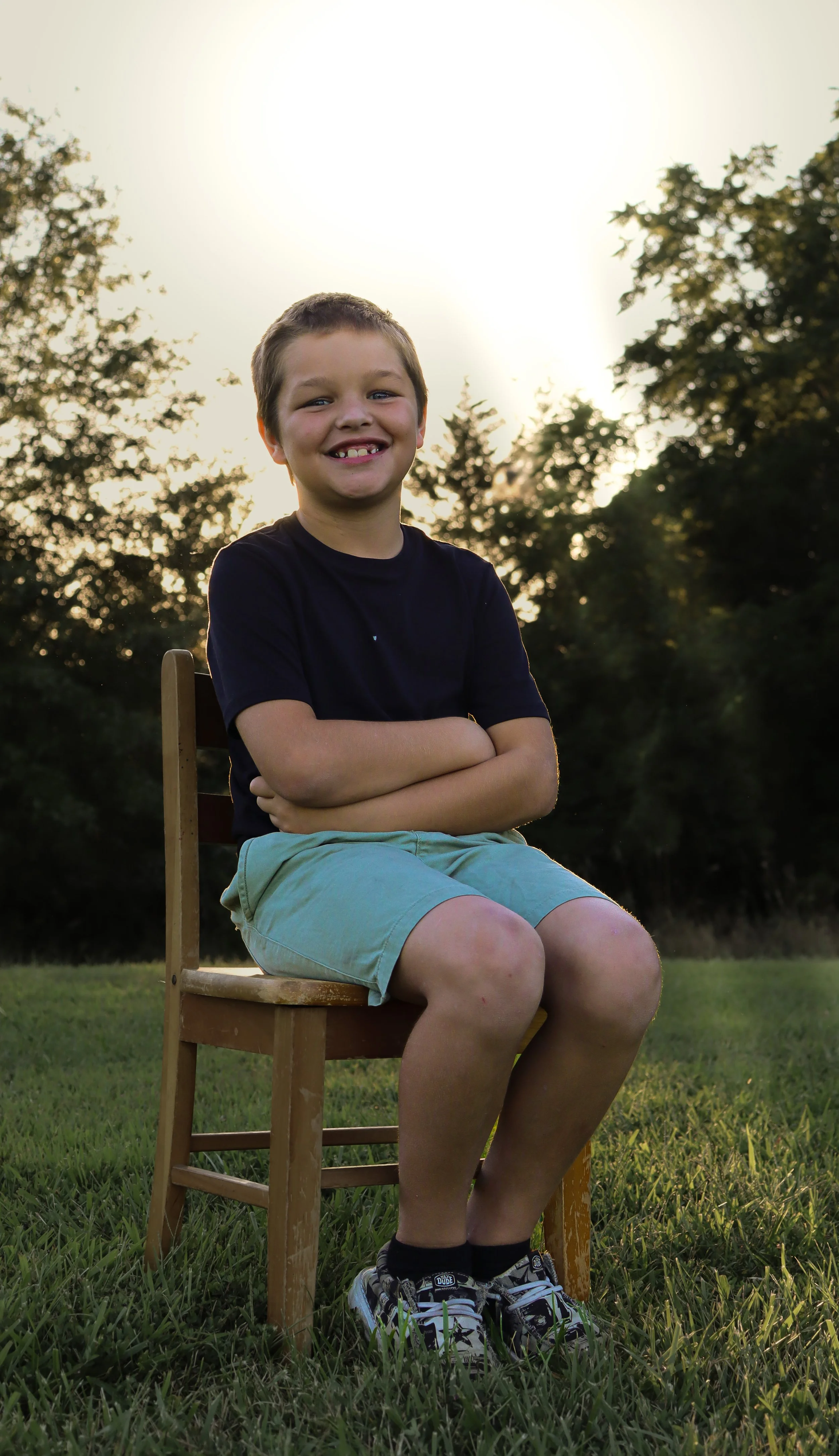 Young boy sitting on a wooden chair outdoors on grass, with trees and sunset in the background, smiling with arms crossed.