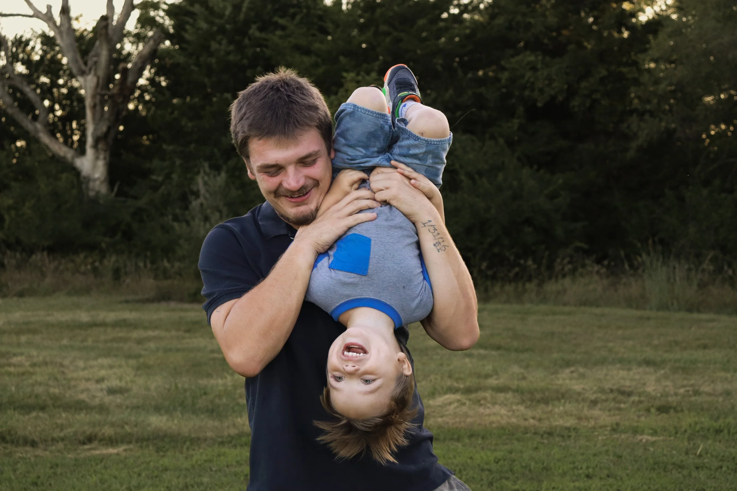 A man swinging a young boy upside down outdoors in a grassy area with trees in the background, both smiling and enjoying the moment.