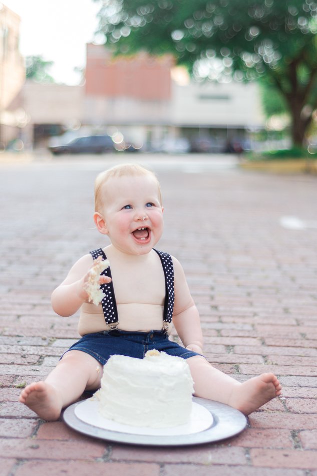 Baby sitting on a brick road eating a white cake, wearing denim shorts and suspenders, with a blurred background of trees and buildings.