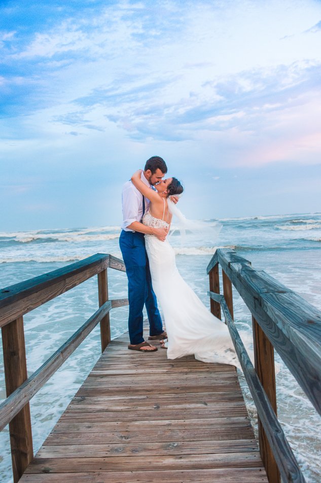 Bride and groom embracing on a wooden pier by the ocean, under a blue sky with clouds.