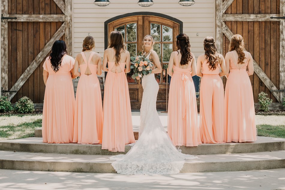 Bride in white gown with bridesmaids in matching pink dresses, standing in front of a rustic barn door, facing the camera.