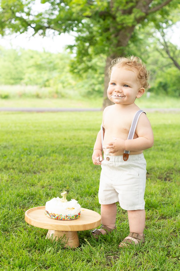 Toddler in overalls outdoors with a small cake on a wooden stand, smiling with frosting on face.
