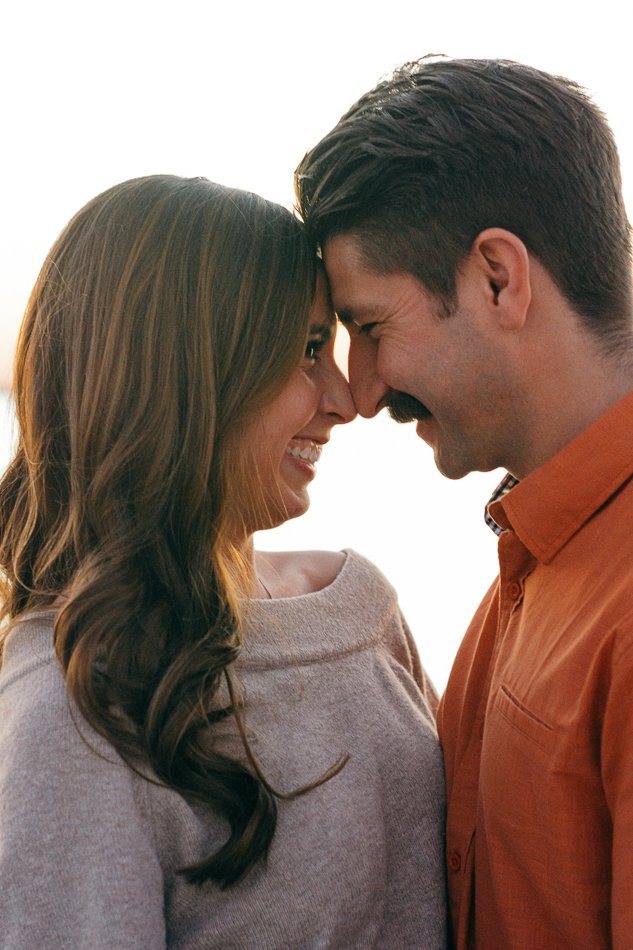 A couple standing close together, smiling and touching their foreheads. The woman wears a gray sweater, and the man is in an orange shirt.