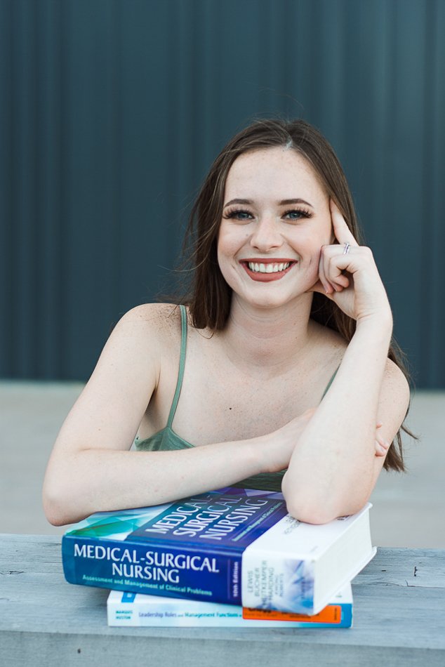 Smiling person with long hair and green top leaning on stack of nursing textbooks.