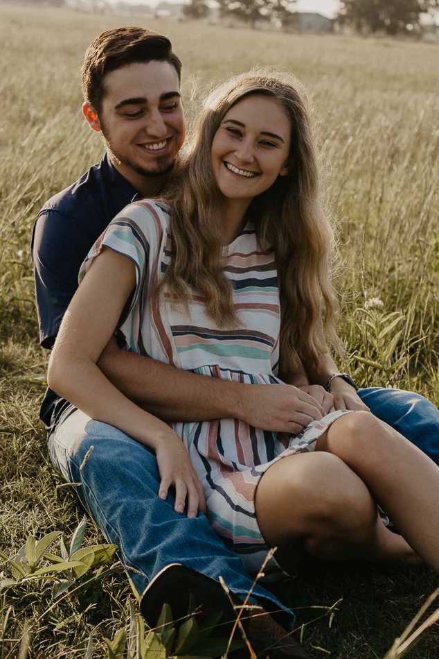 A couple sits in a grassy field, smiling. The woman is in a striped dress and the man is in a dark shirt and jeans.