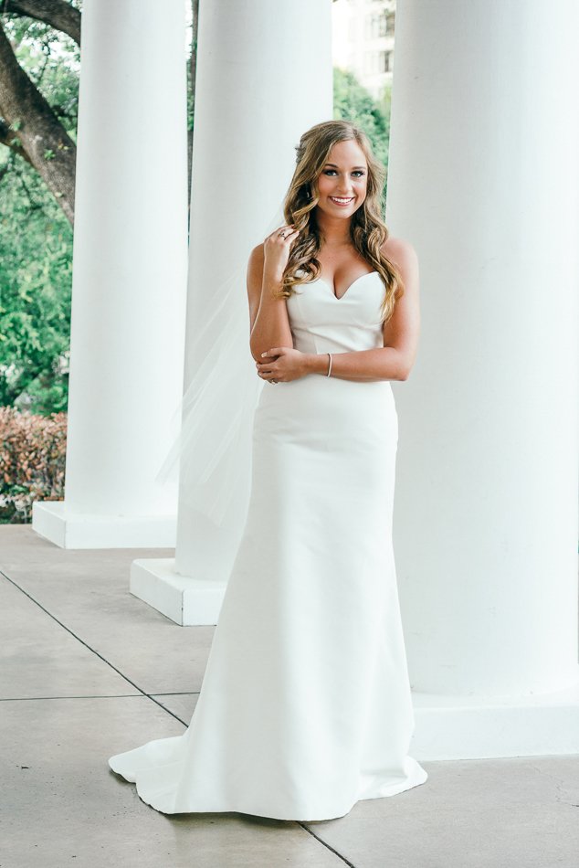 Bride in white gown standing by white columns outdoors in a garden setting.