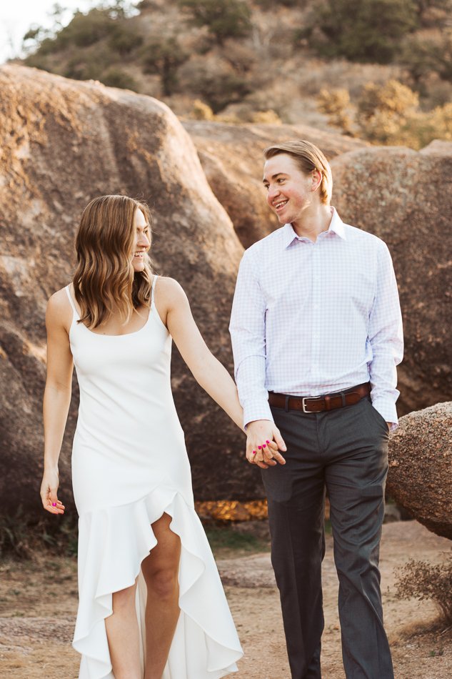 A couple holding hands and smiling, with a rocky outdoor background. The woman is wearing a white dress and the man is wearing a light-colored shirt and dark pants.